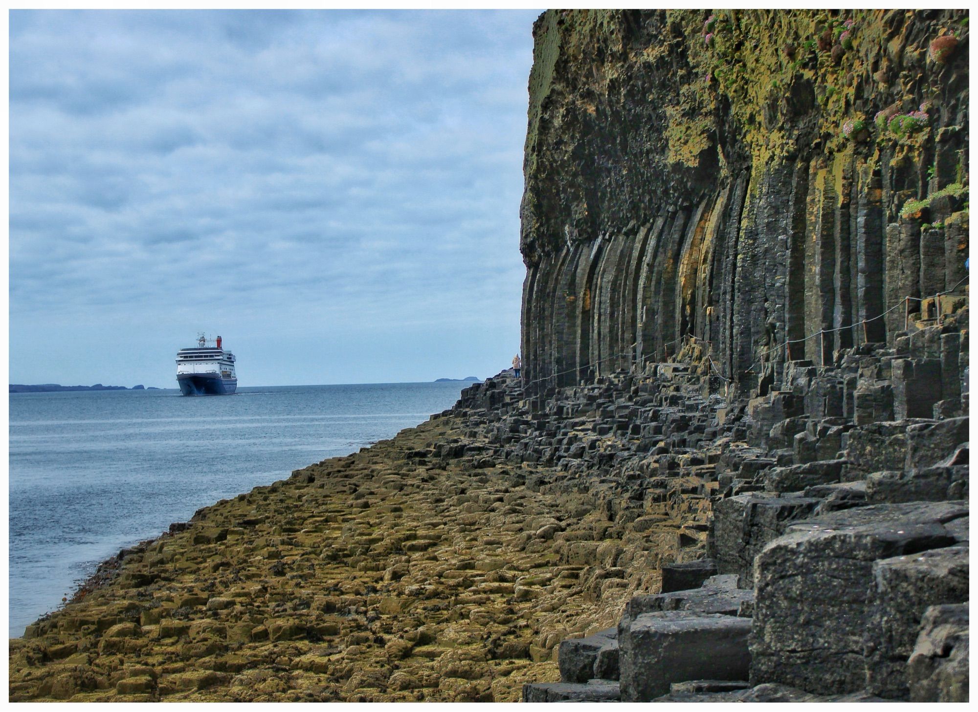 A distant enormous cruise ship sits on the sea next to an island of hexagonal basalt pillars formed from lava flow