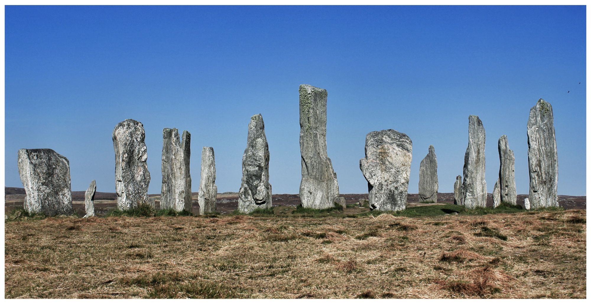 A view of around 15 large standing stones on a grassy piece of moorland against a blue sky.  It’s not possible to tell scale easily from the image, but the monolith at the centre of the stones stands 4.8m. The stones are around 5,000 years old.