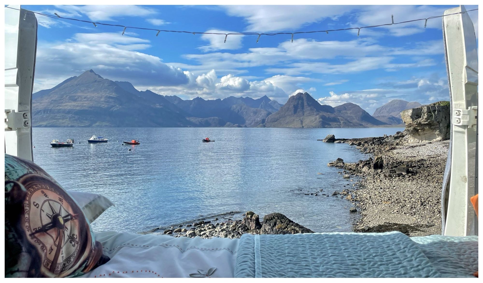 A photograph taken from inside a campervan, looking out across the bed to the view beyond. The foreground shows the bed, with pale turquoise bedding and a cushion with a compass on. The background shows the ridge line of some rocky mountains, set behind a calm sea