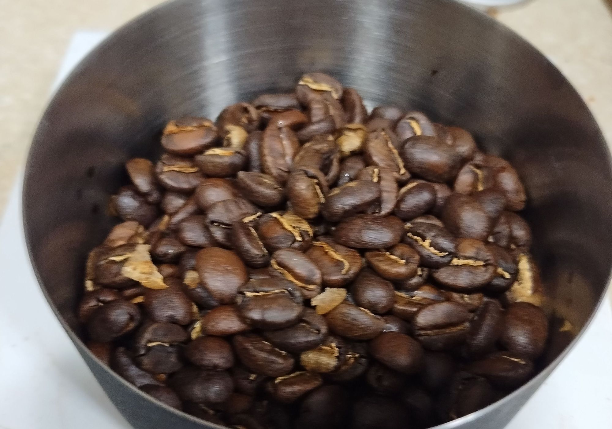 a rounded metal dosing cup with a brushed steel interior and black powder coated exterior holds some roasted Xinabajul coffee beans in it. They're dark brown and the silverskin in their cracks looks yellow. The countertop behind is washed out and just looks white.