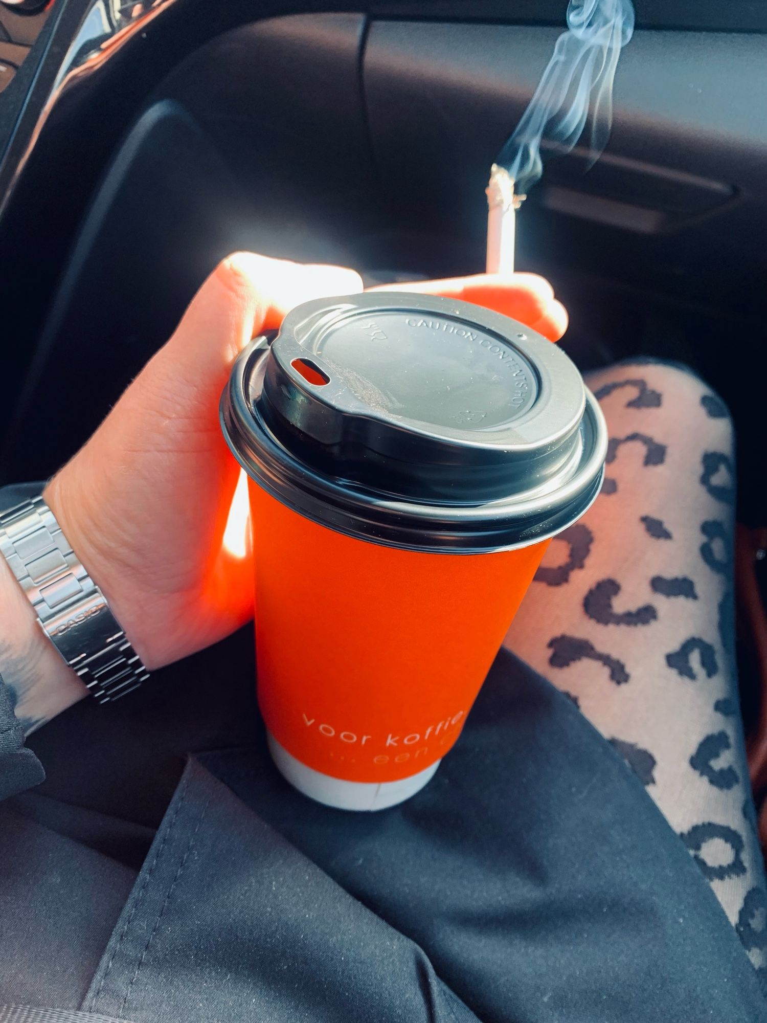 Ich sitze im Auto auf dem Beifahrersitz, man sieht meine Beine und meine linke Hand. Ich trage eine Strumpf mit Leo-Print und einen schwarzen, kurzen Trenchcoat. In meiner Hand halte ich einen Coffee-to-Go Becher und eine brennende Zigarette