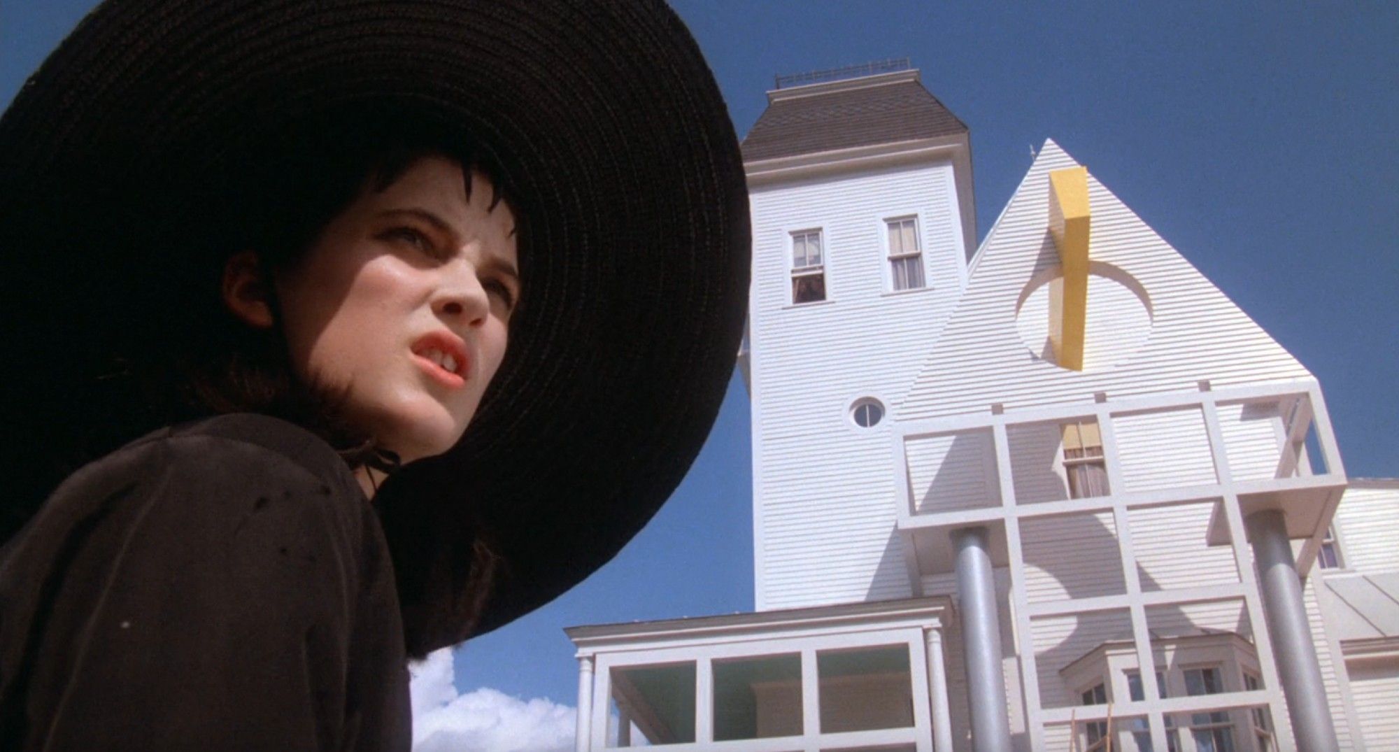 Lydia in daylight outside, wearing all black (including big black wide-brim hat), standing next to the big white house