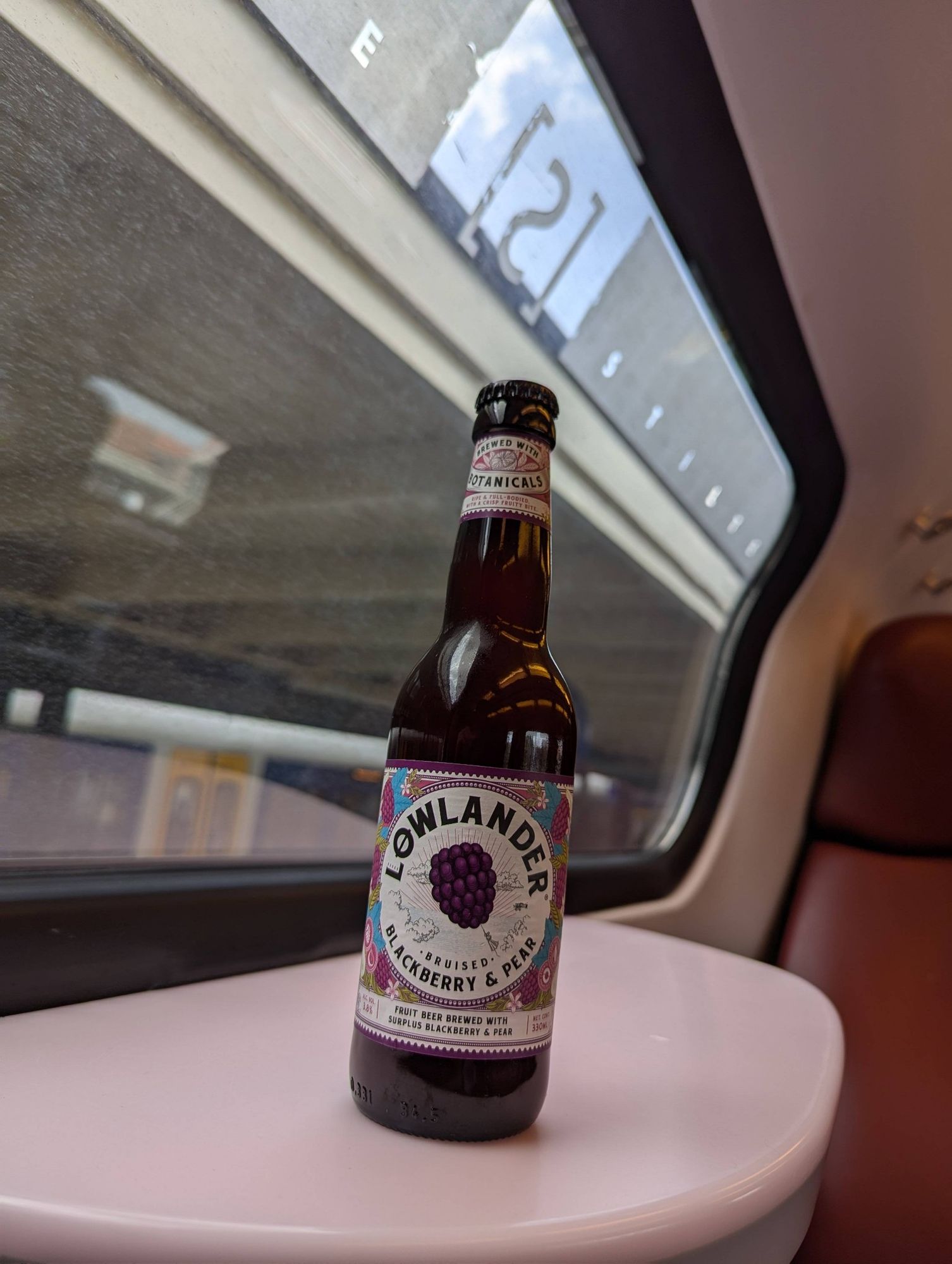 Bottle of lowlander bruised blackberry & pear beer on a table inside a train. Behind the window a train station roof can be seen with a train parked on the other side of the platform