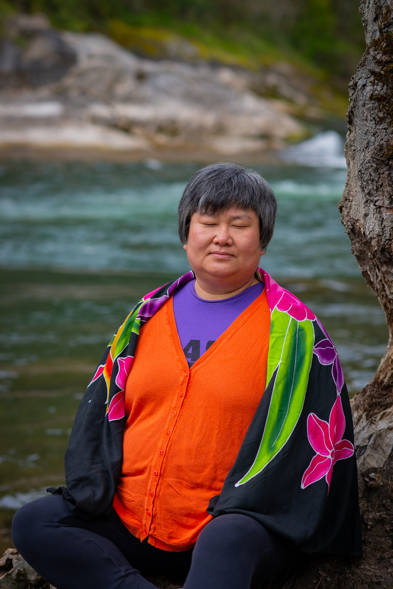 A Chinese-American woman with short gray-black hair meditates by a river, wearing colorful clothing and a flower-patterned shawl.