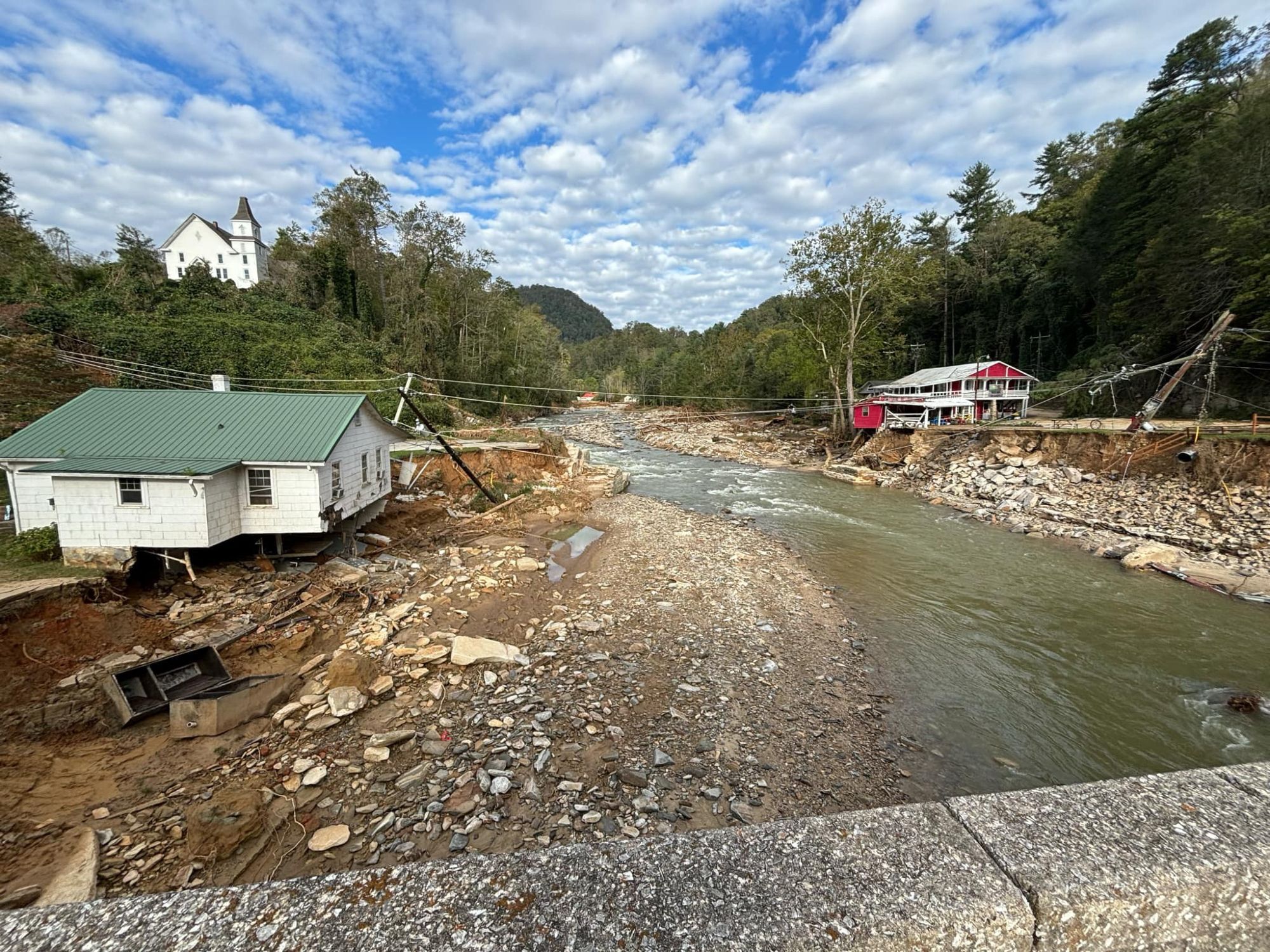 Small houses on either side of a small, shallow river in a mountainous area. On the left bank is a wide bare spot where a house used to be. Seen from a bridge.