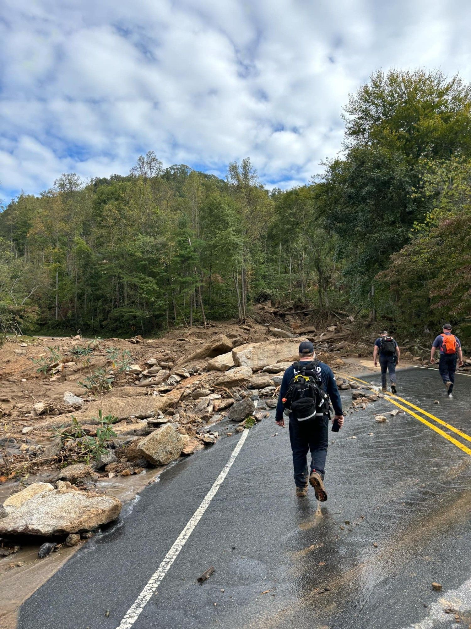 Men with backpacks walking down a wet road in a mountainous area. Ahead of them, the road is covered with boulders and it looks like the road just ends, but it's probably completely covered with debris in the rest of the shot.