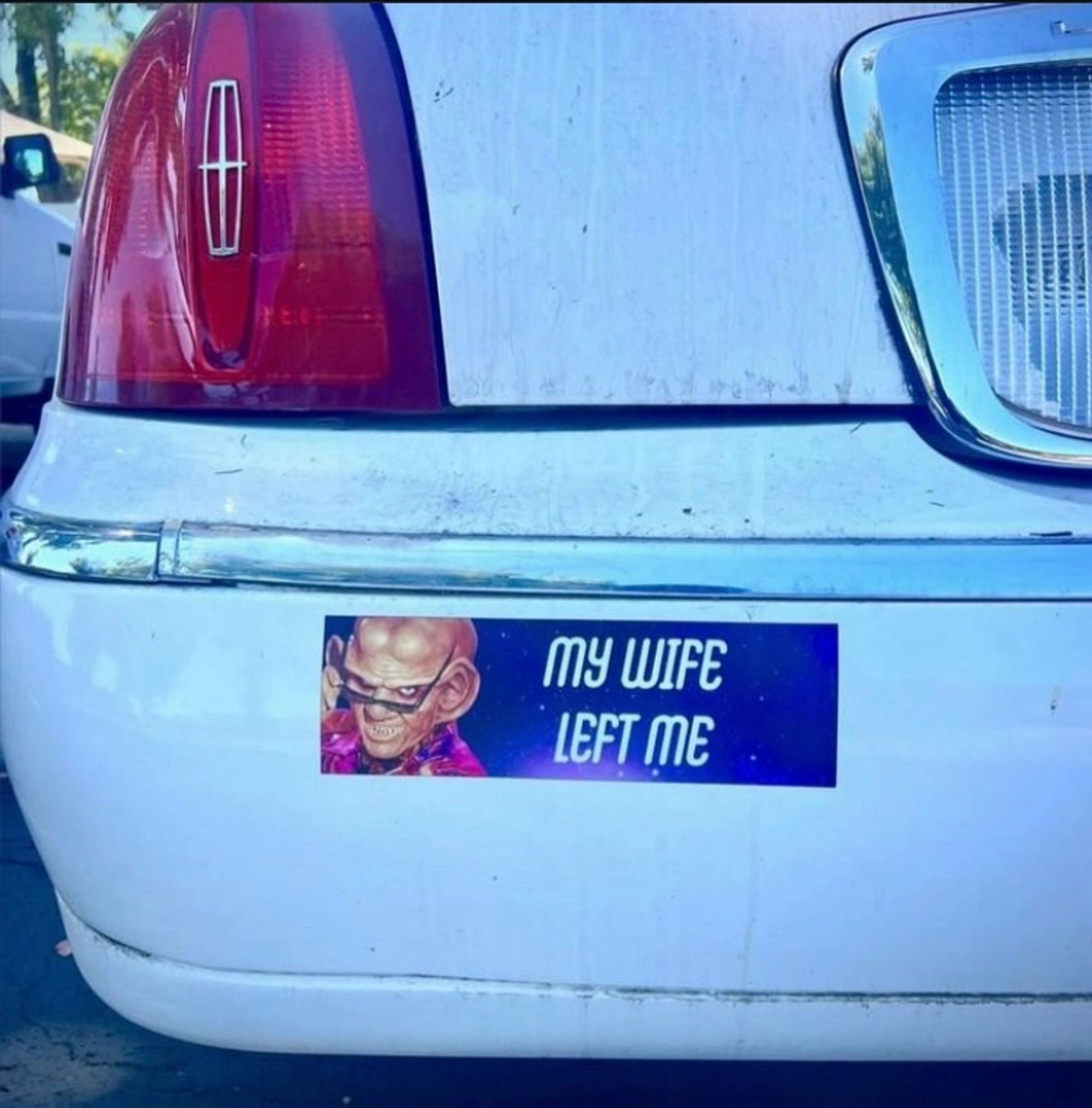 Bumper of a white car with a rectangular sticker featuring a galaxy background and the sexist character Quark from Star Trek: Deep Space Nine pulling down his sunglasses with a smirk. Text in the Star Trek font reads: “My wife left me.”