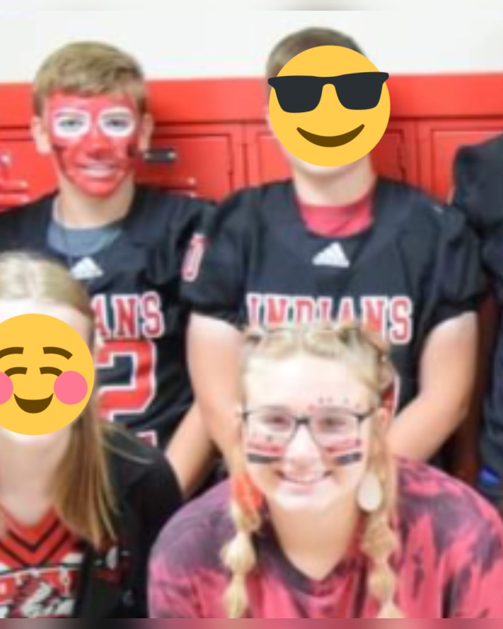 A picture of 4 Tishomingo high school students, two with their faces obscured by emojis. A white presenting boy with blonde hair has his face painted red. A white presenting girl, her blonde hair in braids, has four streaks of paint, two red and two black, reminiscent of war paint, on her cheeks.