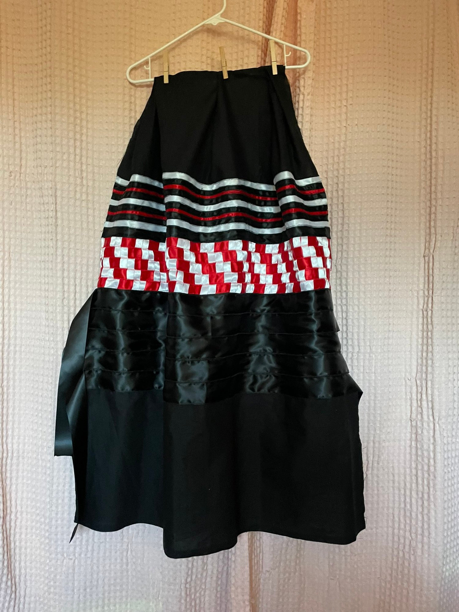 This is the back of the black, white , and red ribbon skirt. It's a black fabric base with alternating thin white, black, and red ribbon near the top that transitions to a white and red ribbon zig zag/stairstep patchwork before fading into 5 black ribbons that have flowy tabs hanging off the side. 