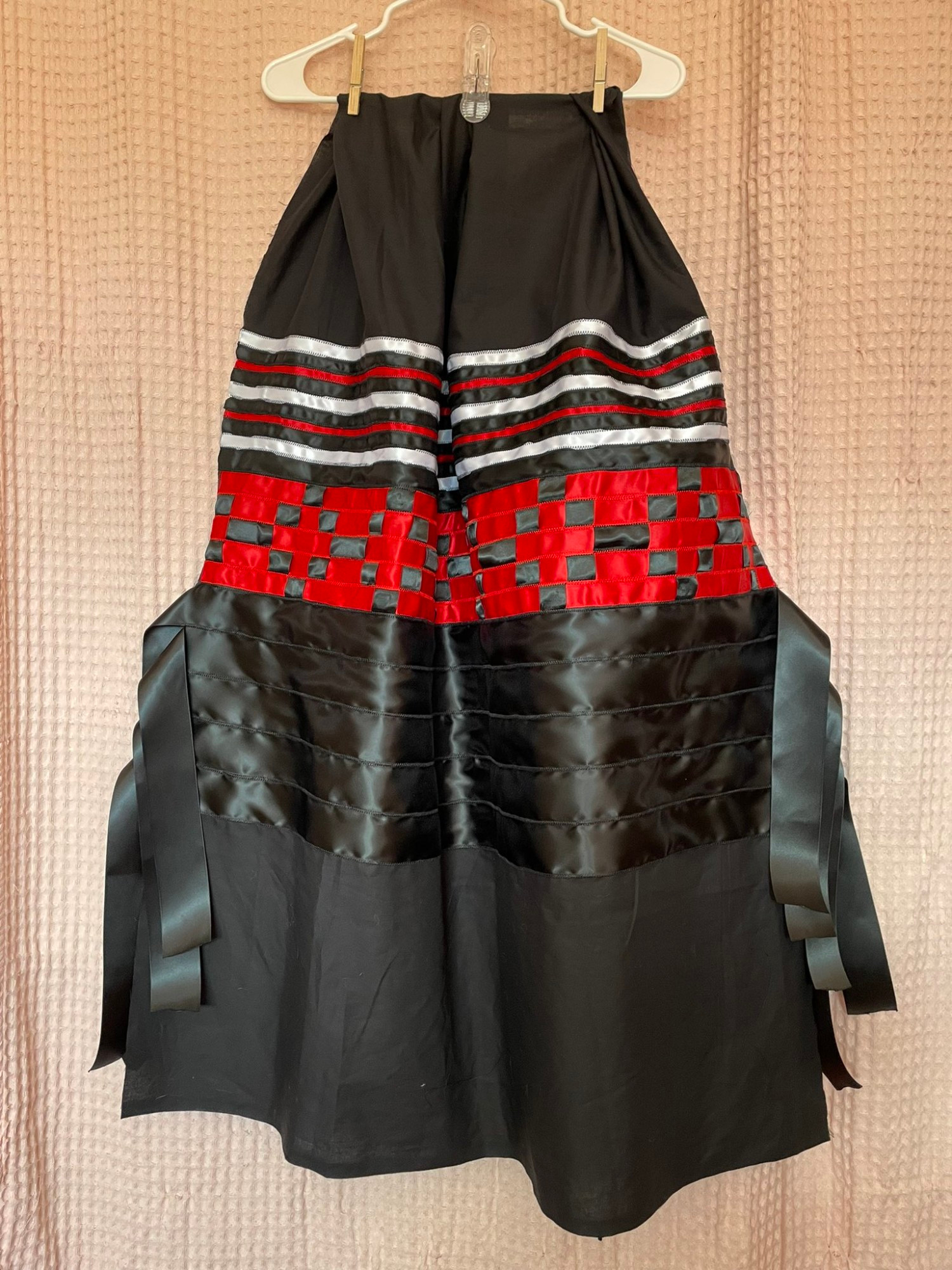 A black, white , and red ribbon skirt. It's a black fabric base with alternating thin white, black, and red ribbon near the top that transitions to a black and red ribbon checkerboard patchwork before fading into 5 black ribbons that have flowy tabs hanging off the side. 