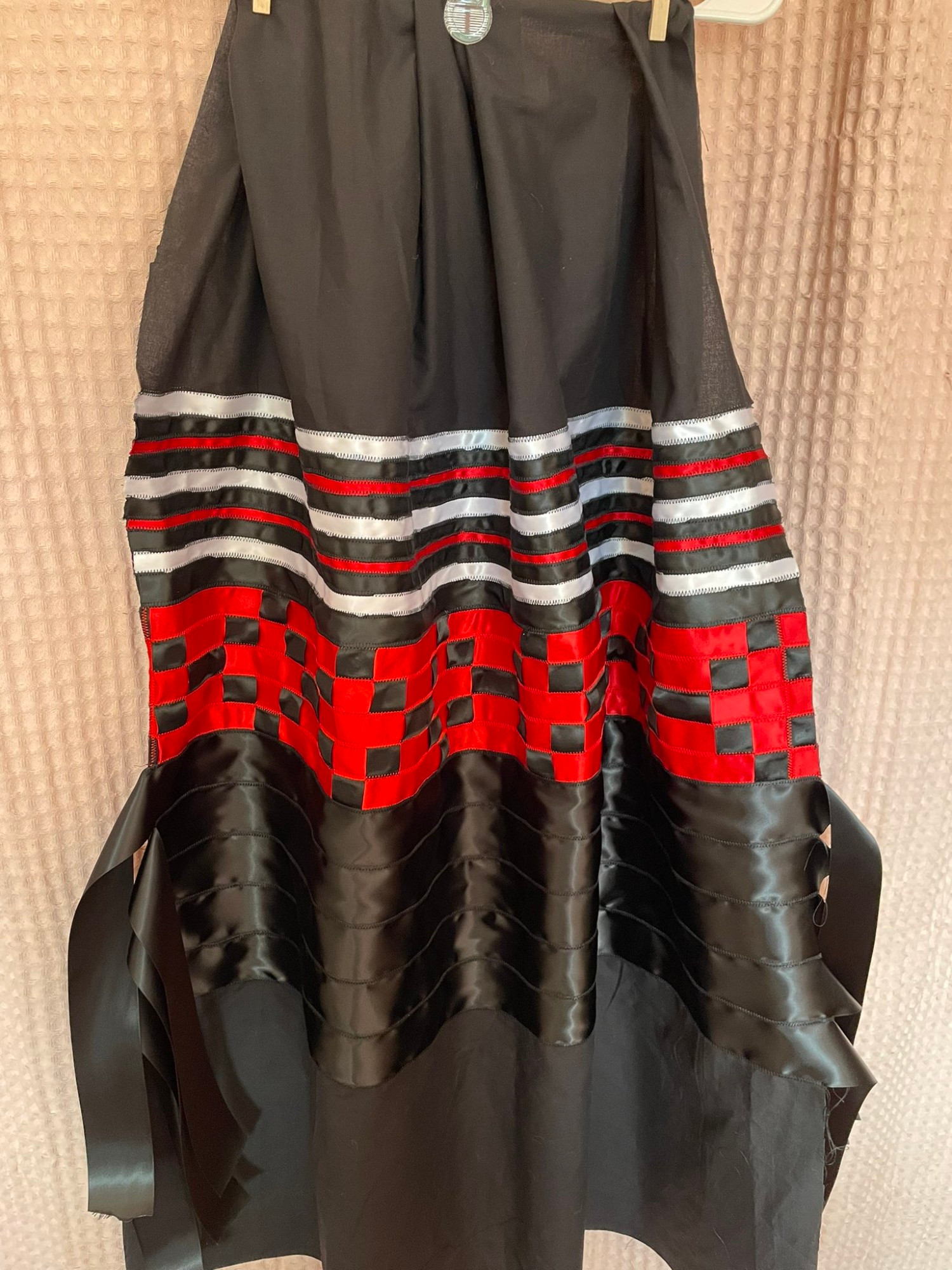 A black, white , and red ribbon skirt. It's a black fabric base with alternating thin white, black, and red ribbon near the top that transitions to a black and red ribbon checkerboard patchwork before fading into 5 black ribbons that have flowy tabs hanging off the side. 