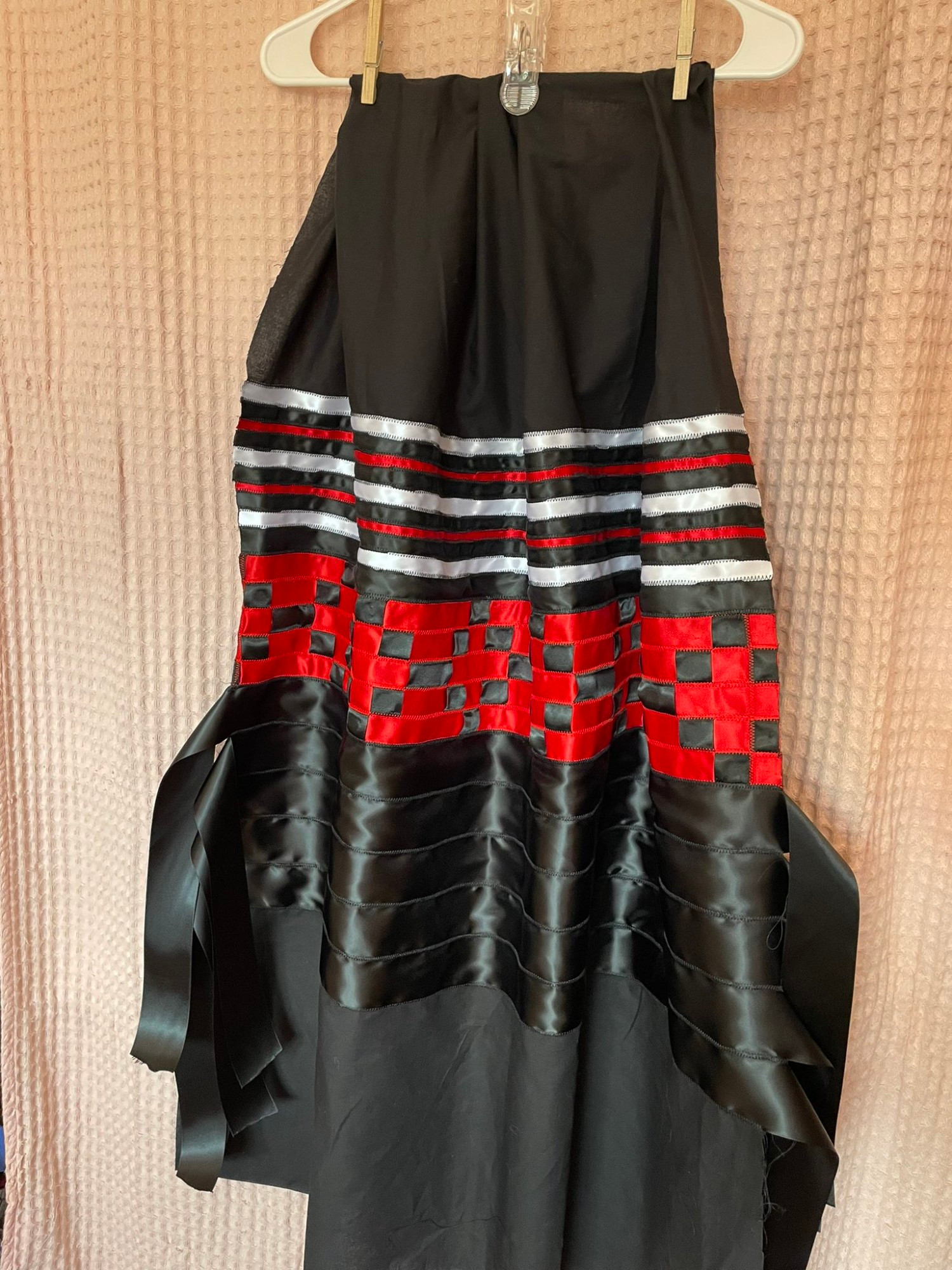 A black, white , and red ribbon skirt. It's a black fabric base with alternating thin white, black, and red ribbon near the top that transitions to a black and red ribbon checkerboard patchwork before fading into 5 black ribbons that have flowy tabs hanging off the side. 