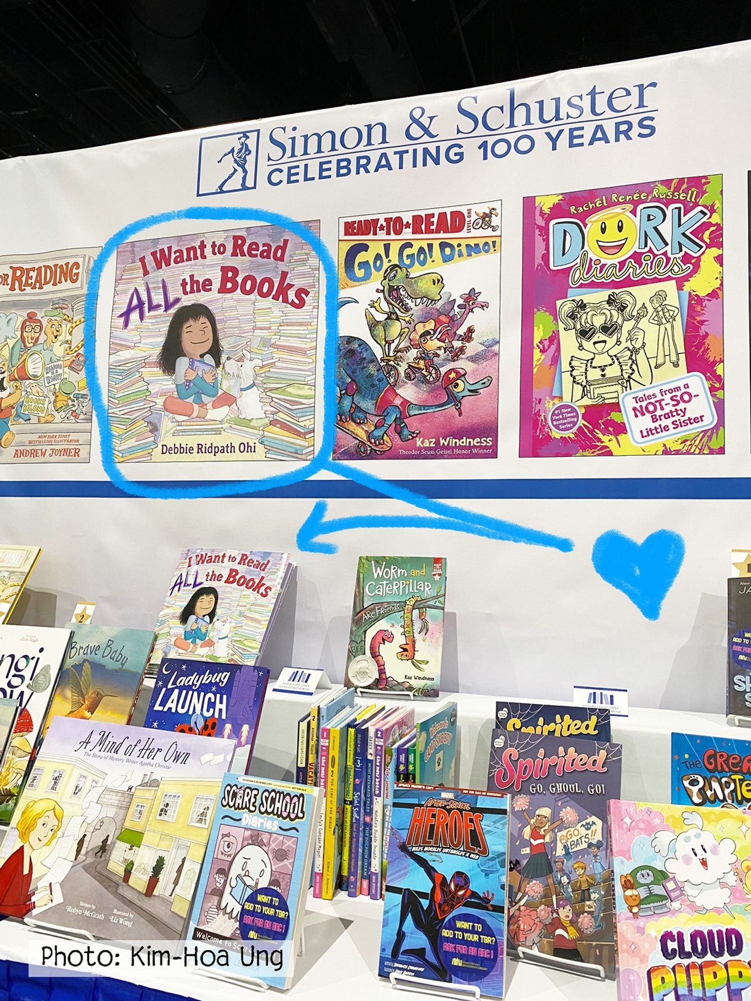 Display booth for Simon & Schuster, with a banner saying they are celebrating 100 years. Several children's books are showcased. One book, "I Want to Read ALL the Books" by Debbie Ridpath Ohi, is highlighted with a blue circle and an arrow pointing to another display of the same book below. Other books in the display include "Go! Go! Dino!" by Kaz Windness and "Dork Diaries" by Rachel Renée Russell. The photograph is credited to Kim-Hoa Ung. #kidlit