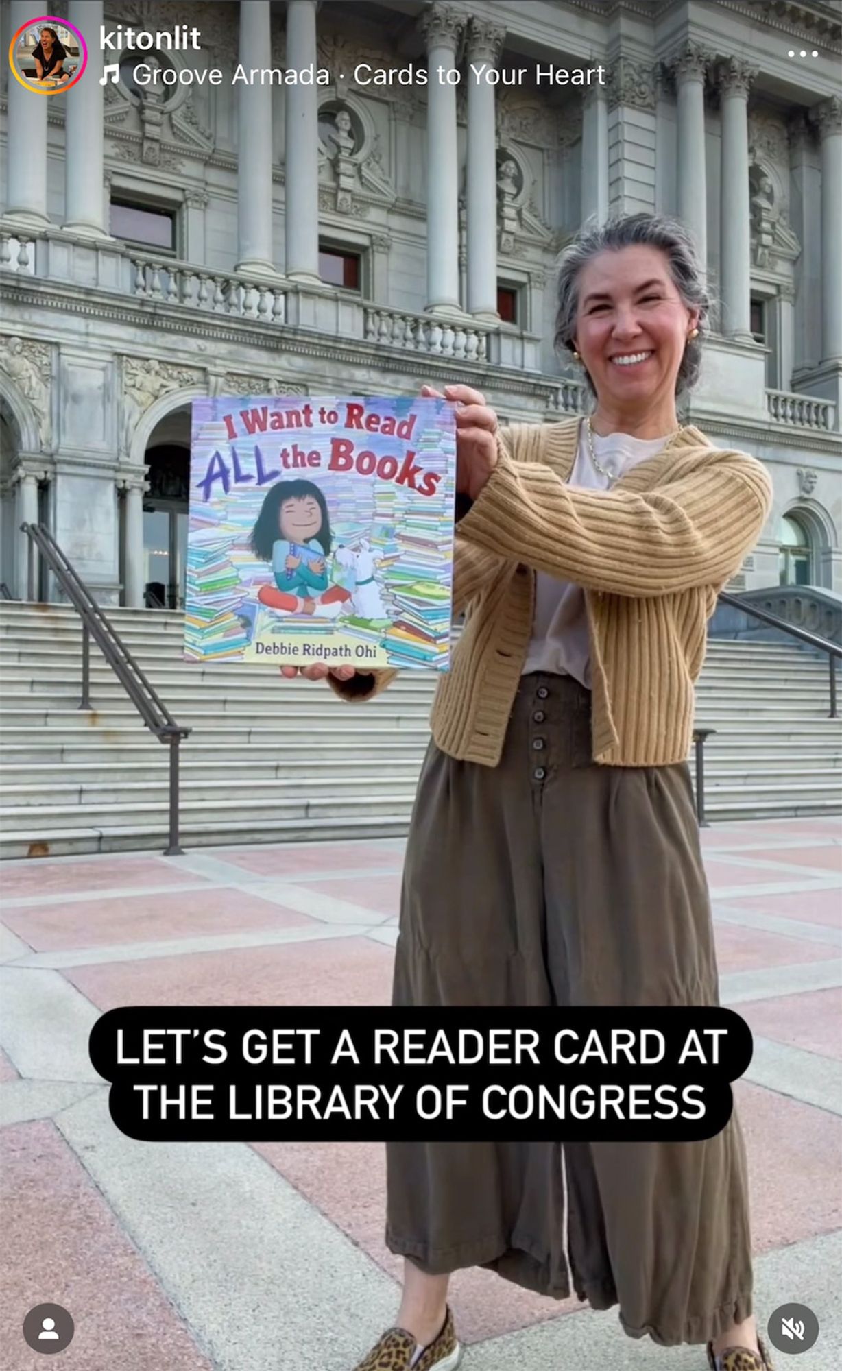 A smiling woman holds the children's book I Want to Read All the Books by Debbie Ridpath Ohi in front of the Library of Congress. Text at the bottom reads, "Let's get a reader card at the Library of Congress." The video suggests excitement about reading and encourages library participation, emphasizing access to books and knowledge.