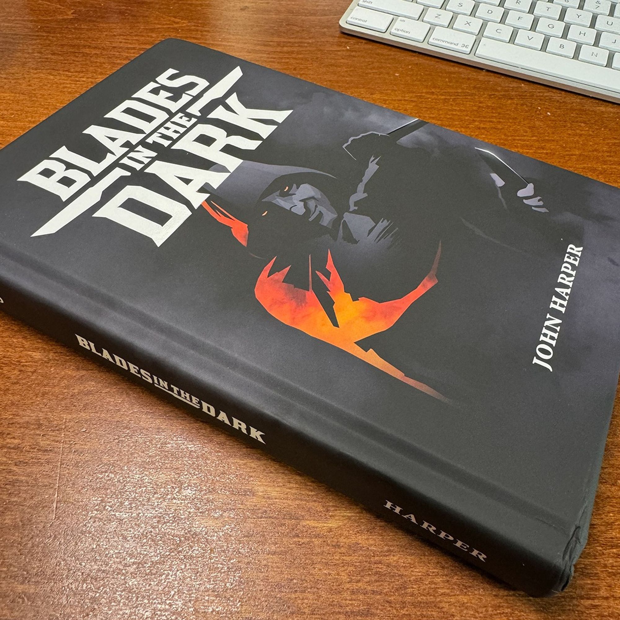 A hardcover copy of the book Blades in the Dark by John Harper rests on a wooden desk. The book's cover features a dark, shadowy figure with a hood, accented by a bold orange-red flame design. A white keyboard is partially visible in the background.