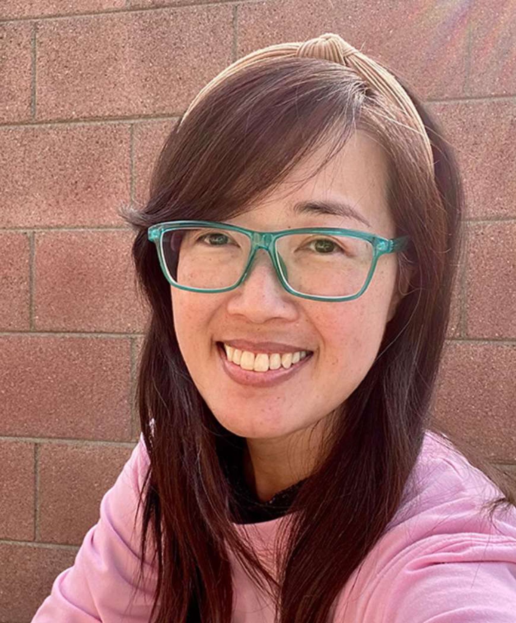 Headshot photo of a young smiling Asian woman with dark hair and a brown headband, wearing turquoise glasses and a pink shirt.