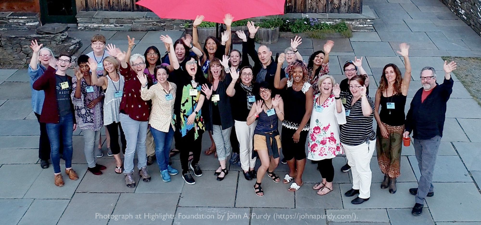 A group of people stand outdoors on a stone patio, smiling and waving enthusiastically at the camera. The group includes a diverse mix of individuals, all appearing joyful and engaged. A red umbrella is visible in the background. The caption at the bottom credits the photograph to John A. Purdy at the Highlights Foundation.