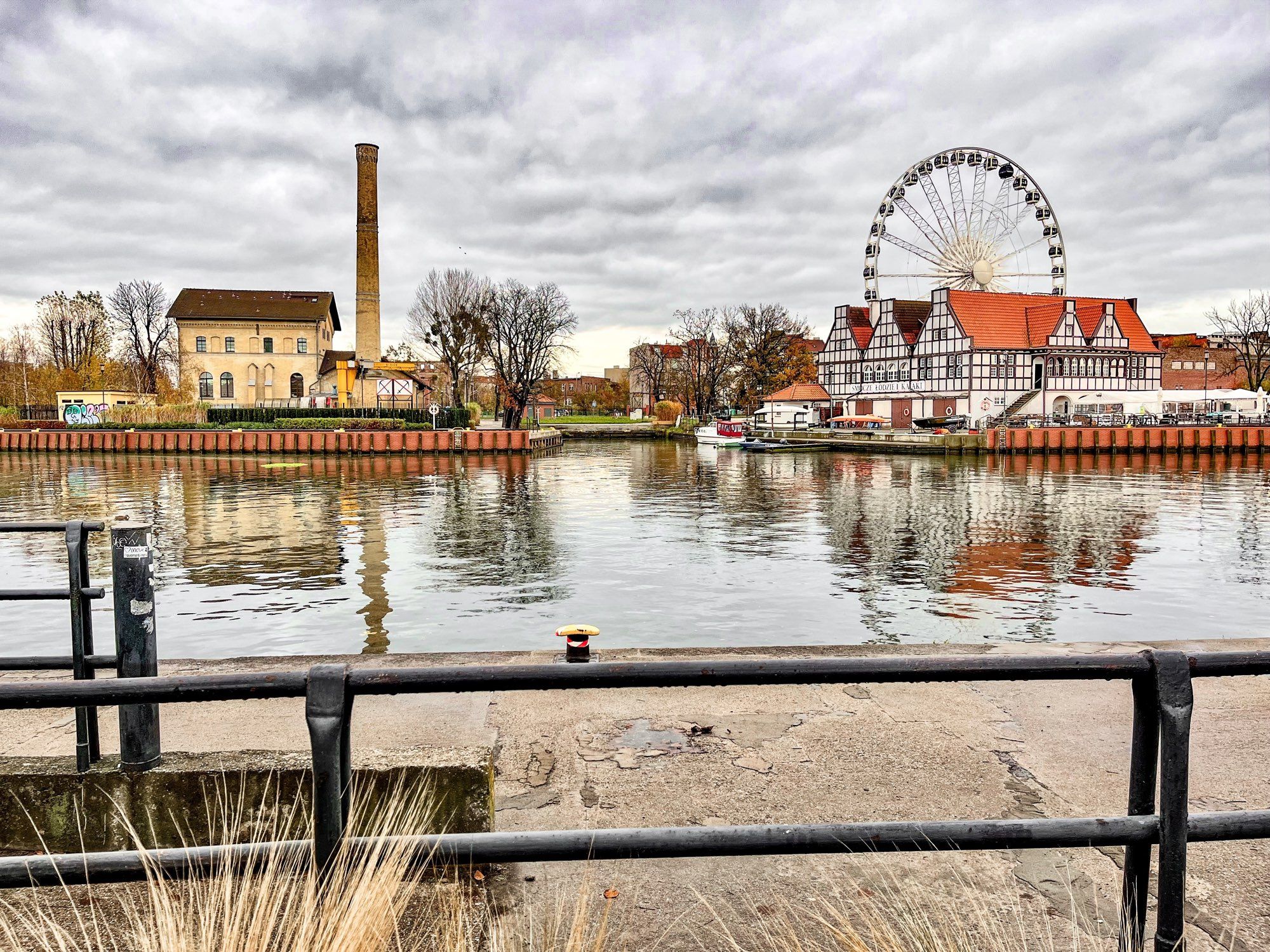 Blick über eine recht glatte Wasseroberfläche auf ein verlassen wirkendes Gebäude auf der linken und - neben einer kleineren Einmündung - einem größeren Fachwerkbau mit roten Dachflächen auf der rechten Seite, dieser wird von einem mittelgroßen Riesenrad überragt. Der mit grauen Wolkenstreifen überzogene Himmel spiegelt sich mit den Gebäuden und einigen Bäumen im Wasser, im Vordergrund eine verwitterte Kaimauer aus Beton mit einer Sitzbank (links) und ganz vorne einem Geländer aus Stahlrohren.