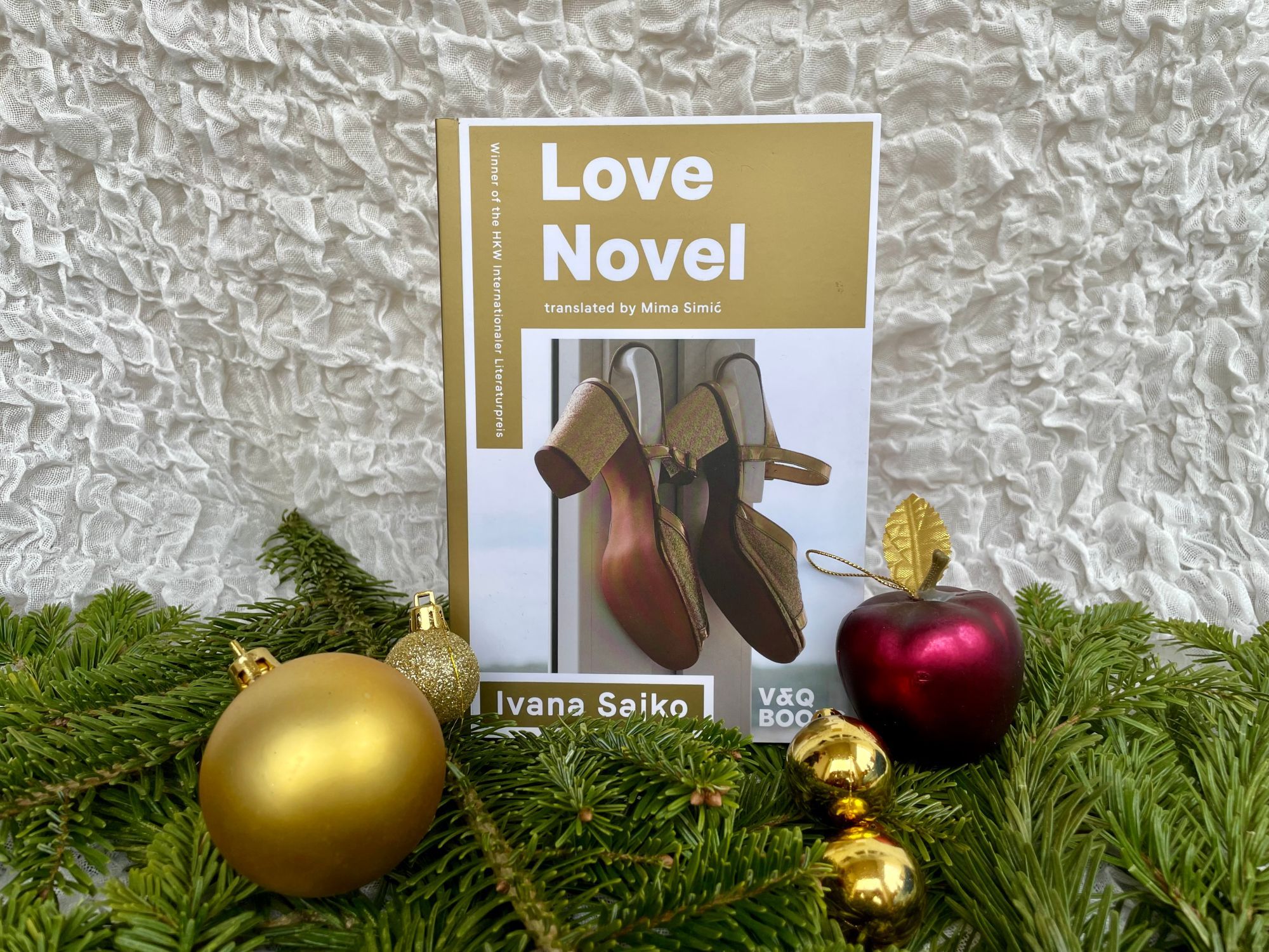 Our golden book LOVE NOVEL with golden high heels on the cover, bedded on pine branches and nestled with golden baubles and a red plastic apple (with a golden leaf), against a curly white background used in an attempt to simulate snow.