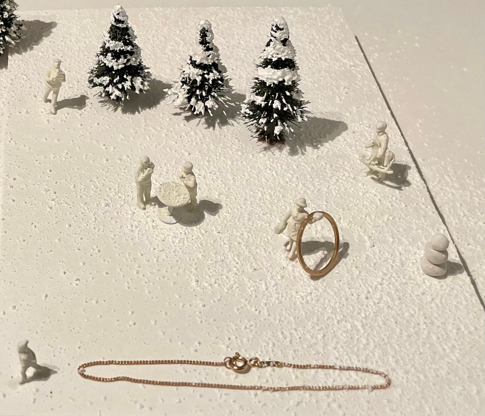 Teeny tiny featureless white figurines and teeny tiny fir trees splatted with possibly Tippex to simulate snow. One of the figurines is balancing a golden hoop earring over its outstretched, which is about the same height as the figurine itself.