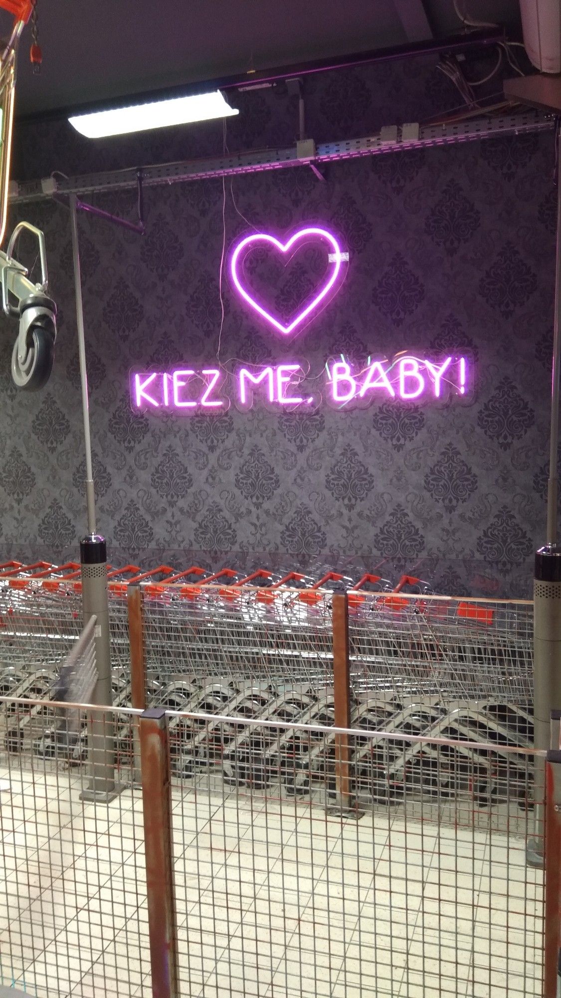 Einkaufswagen im Supermarkt.
Darüber pinkfarbenes Neonherz mit gleichfarbigen Schriftzug "KIEZ ME. BABY!