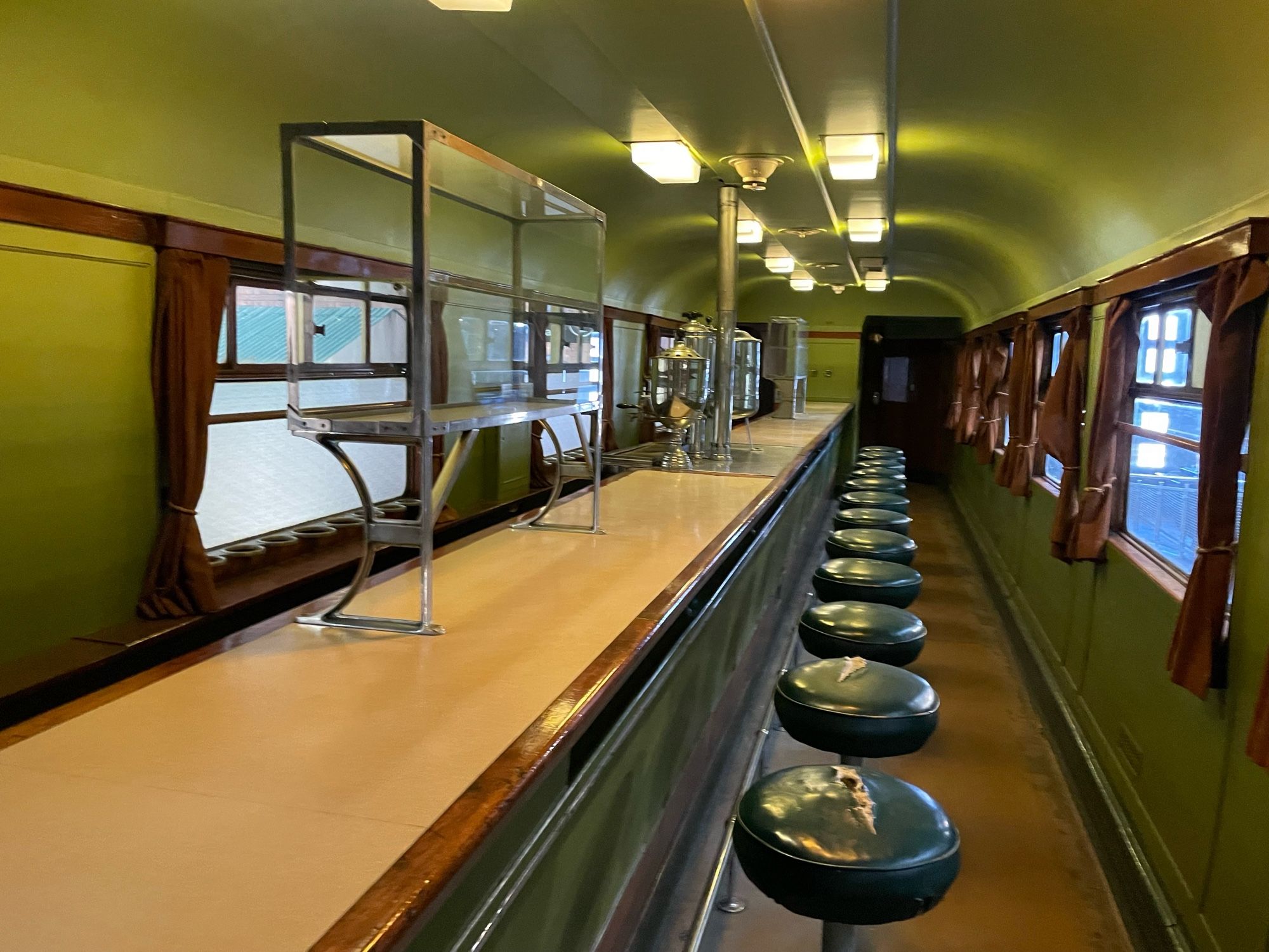 Swindon STEAM Great Western Railway/God’s Wonderful Railway bar in a buffet carriage with green topped bar stools, wooden counter running down the whole carriage. Counter edged with dark wood and red curtains at the windows.