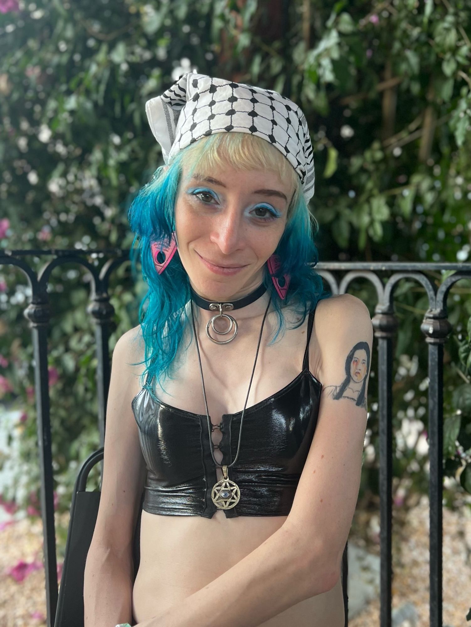 Jan with blue dip-dyed hair and blue eye shadow sitting in front of green bushes and wearing a white keffiyeh bandanna, pink earrings and vinyl black crop top