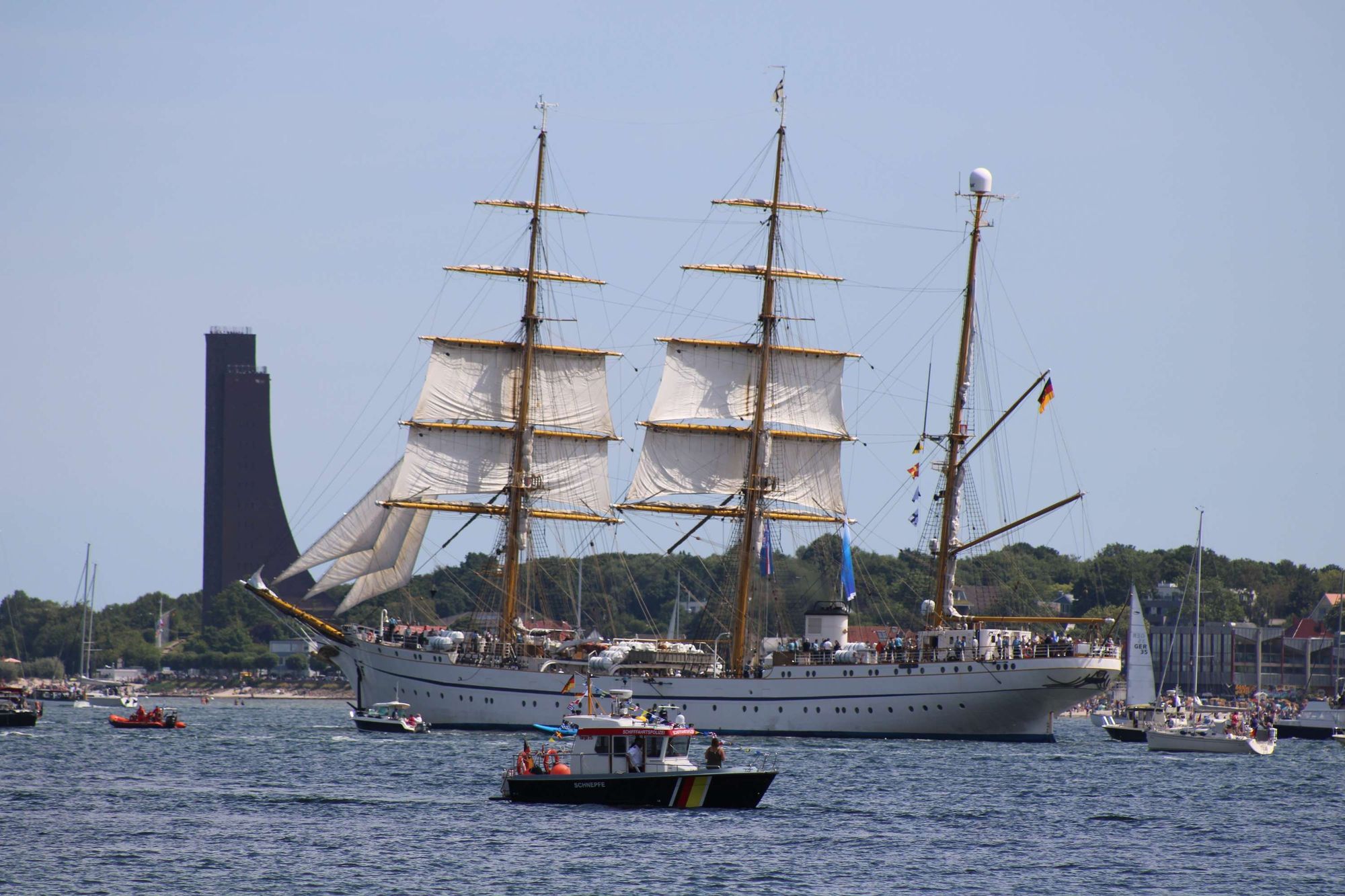 Die Gorch Fock führt die Winjammerparade 2024 an. Sie segelt aus der Kieler Förde heraus. Im Hintergrund sieht man das Marine-Ehrenmal in Laboe.