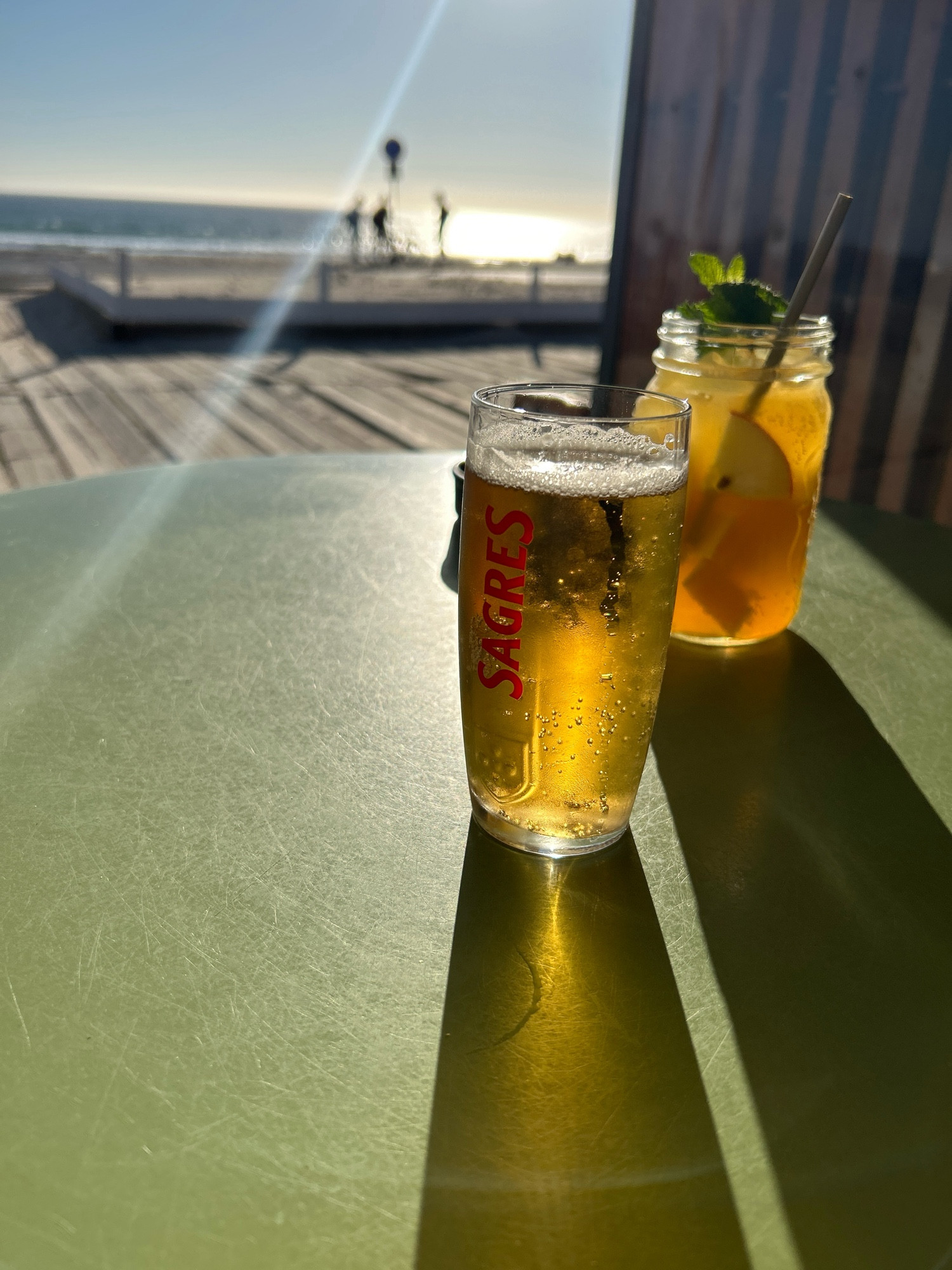 Bier und weißer Sangria auf einem grünen Tisch. Im Hintergrund eine Terrasse, Promenade, Strand und Meer, ich welchem I h die Sonne spiegelt.