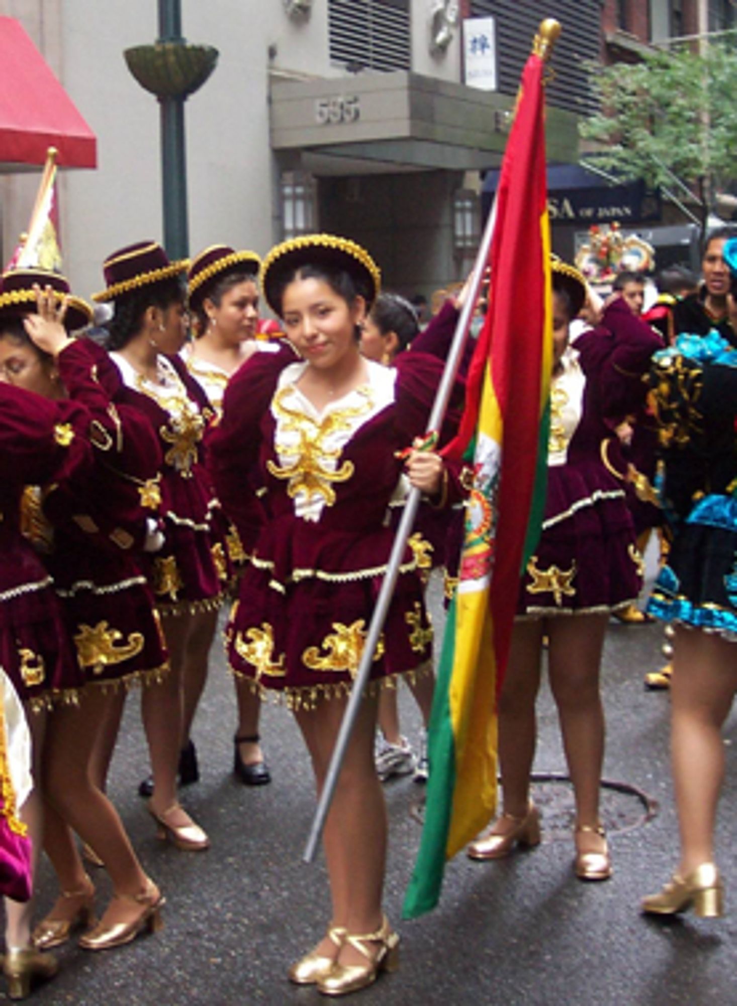 Photo of Aida (center) in a Bolivian folkloric dance costume, a red velvet high shouldered top, short pleated skirt with white and gold accents. Holding a Bolivian flag.