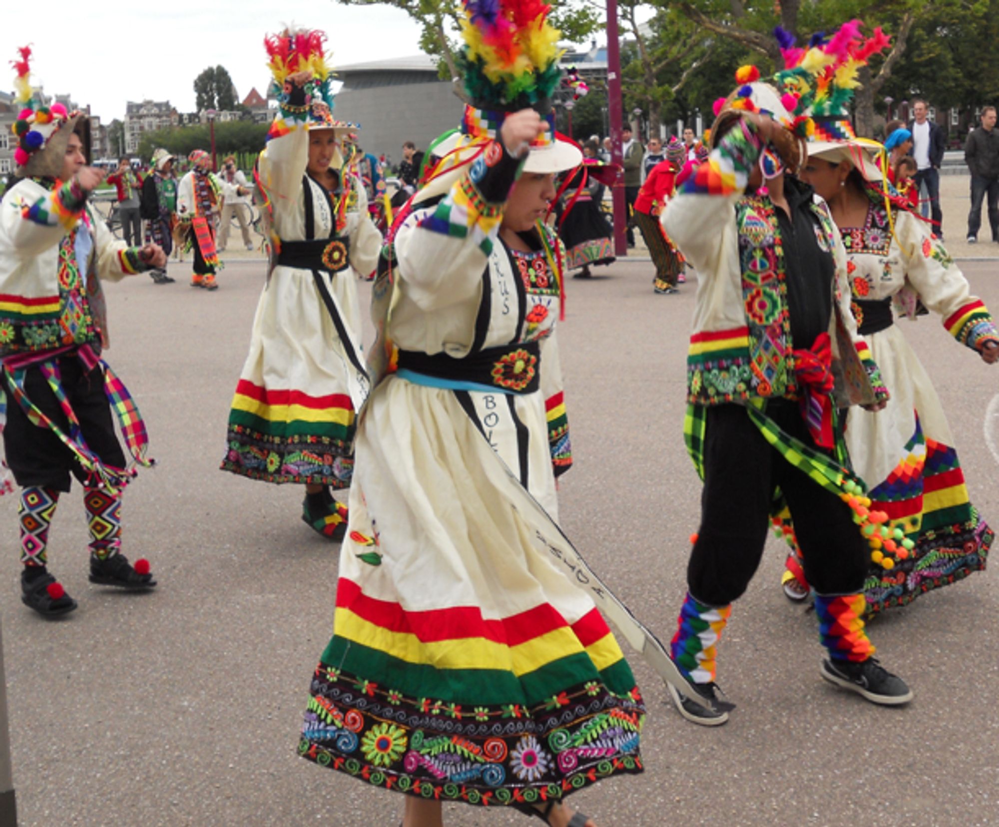 Photo of Aida (center) in a folkloric Tinku costume. A long-sleeved white woolen ankle-length dress. Very colorful embroidery, borders, covered in ribbons, neon feathers sticking straight up from a brimmed woolen hat.