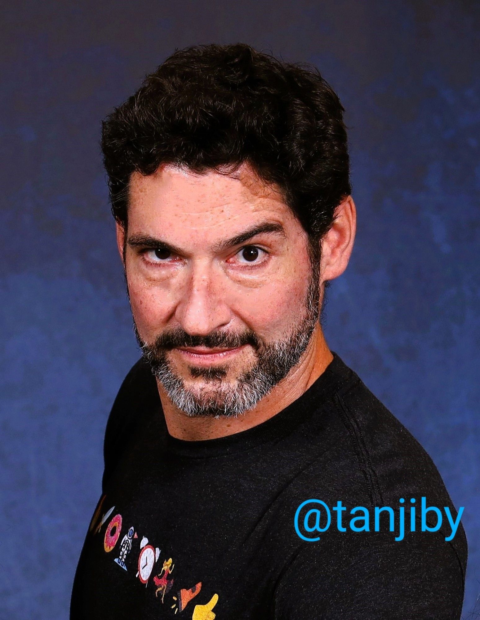 Tom Ellis at MagicCon 2023 - he's wearing a black shirt with the emojis of sending Mom home. His dark hair's curly, salt+pepper beard, no glasses. He's looking "develish" into the cam, one eyebrow raised