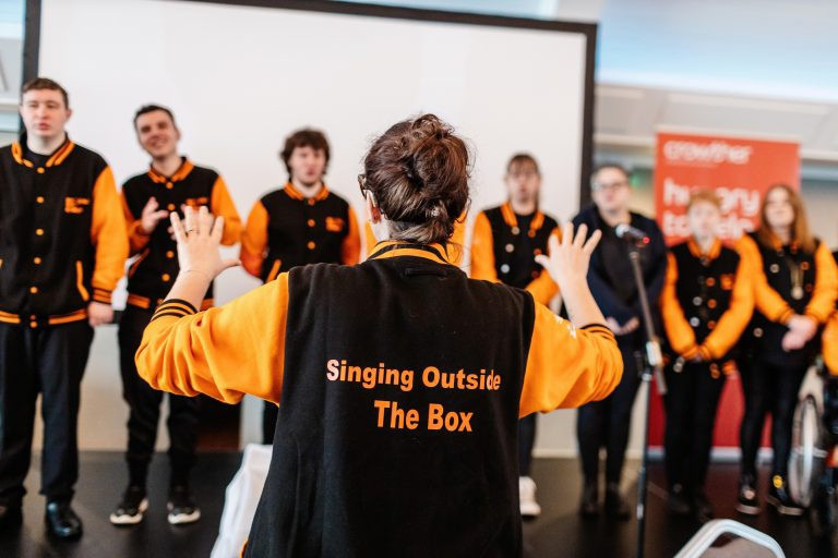 A group of young adults are singing in a choir. In the foreground they are conducted by a choir leader. On the back of her jacket it says "singing outside the box."