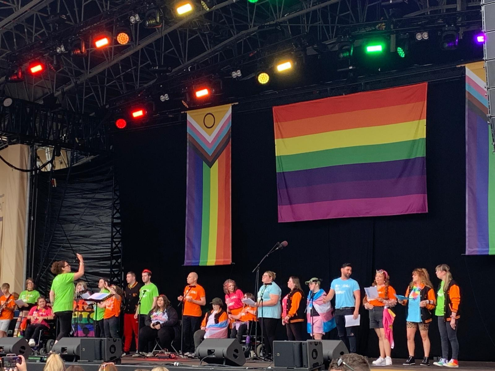 A group of 21 people in colorful tshirts are in a line on a large stage. Some are standing, some are seated in wheelchairs. They are singing. Hanging behind them are large pride flags.