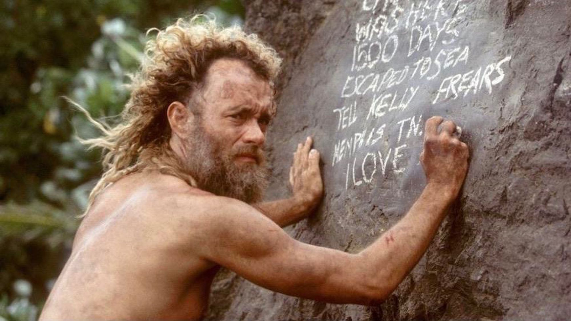 A shirtless Tom Hanks looks wistfully out to sea (idk I’ve never actually seen the movie) in the film Castaway. He is using a rock to write a note on a rock. He looks haggard af and has no clothes 