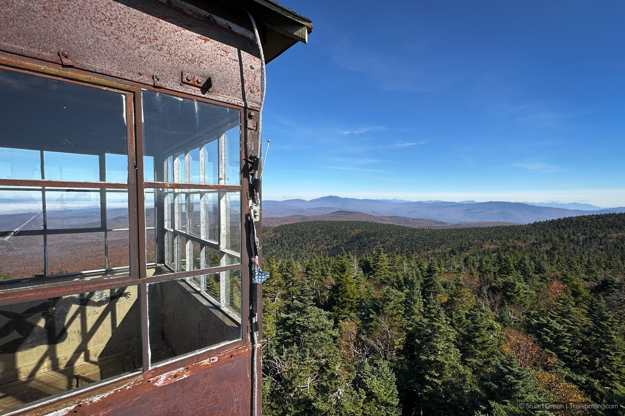 A high vantage point view of a dense forest with varying shades of green, extending towards rolling hills and distant mountains under a clear blue sky. Part of the weathered fire tower cabin from where the photo is taken is visible on the left, featuring old window frames with some broken panes.