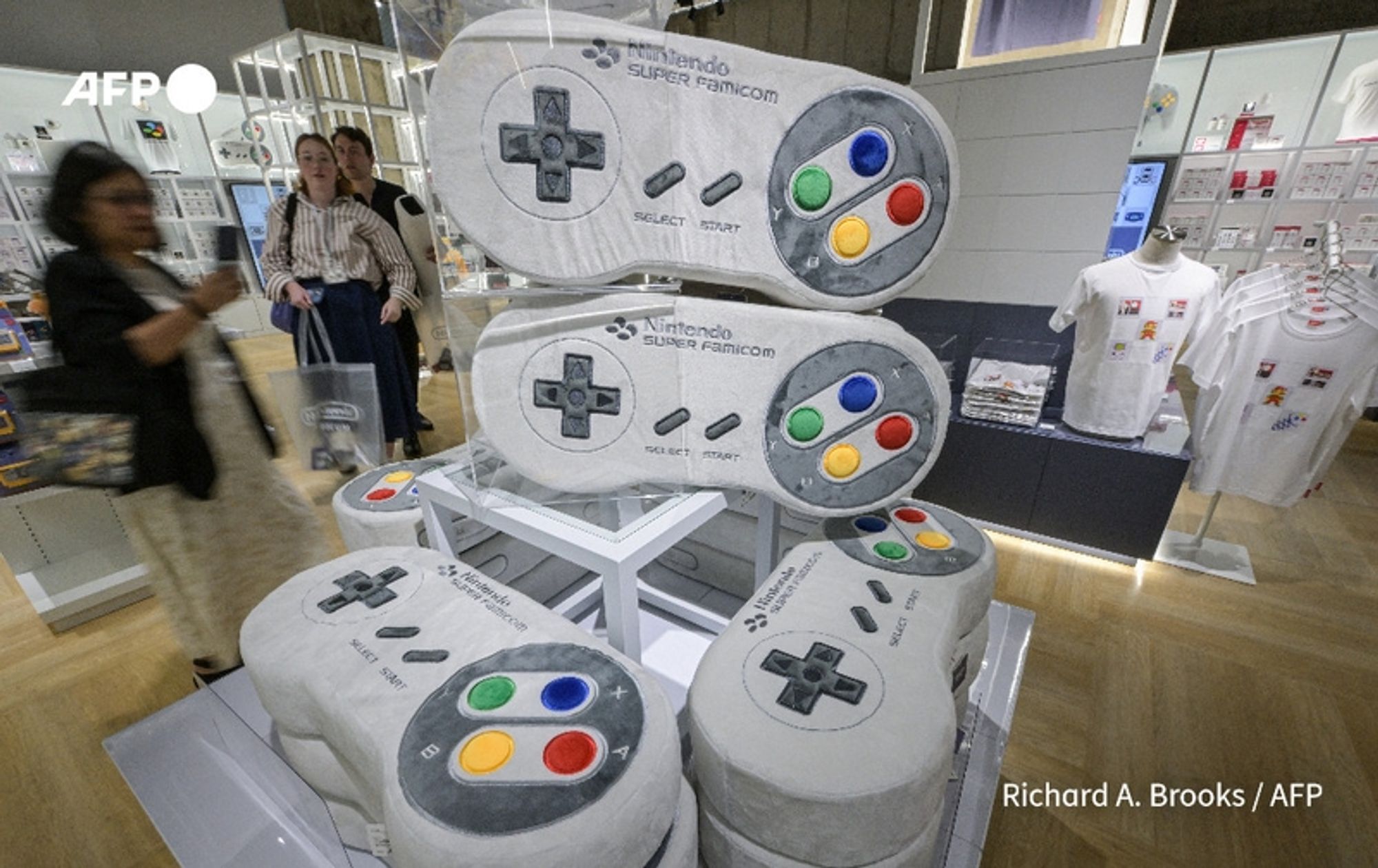 A display inside the Nintendo museum in Kyoto, featuring oversized, stylized sculptures of the Nintendo Super Famicom controllers. The controllers are arranged in a stack, prominently showcasing their distinct design, which includes the classic directional buttons, along with colorful action buttons: red, yellow, blue, and green. Surrounding the display, a few visitors can be seen, including a woman with dark hair wearing a light-colored dress and holding shopping bags, and others in stripes walking behind her. In the background, shelves filled with Nintendo merchandise, including T-shirts and other memorabilia, are visible, emphasizing the museum's focus on the history and evolution of the brand from its origins in playing cards to video games. The overall atmosphere is lively, reflecting the nostalgia and excitement of Nintendo's rich gaming legacy.