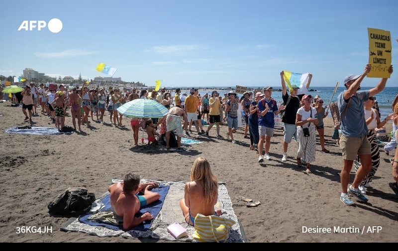 A lively beach scene filled with demonstrators gathered on a sandy shoreline under a clear blue sky. In the foreground, a woman sits on a beach towel, wearing a yellow bikini, with her back to the viewer, while a man beside her lies on a towel in shorts. Both appear relaxed, but the crowd behind them is animated. 

A diverse group of protesters, some holding colorful flags and signs, marches along the beach, demanding restrictions on tourism in the Canary Islands. The signs feature messages such as "Canarias tiene un límite," highlighting their concerns. Some participants wear casual summer clothing, including shorts, tank tops, and hats, while others carry umbrellas for shade. 

The beach is busy, with more people in the background, some sunbathing, while others engage in conversation or join the protest. The atmosphere is energetic, filled with a sense of urgency and community as people advocate for their cause, all set against the backdrop of the sparkling Atlantic Ocean.