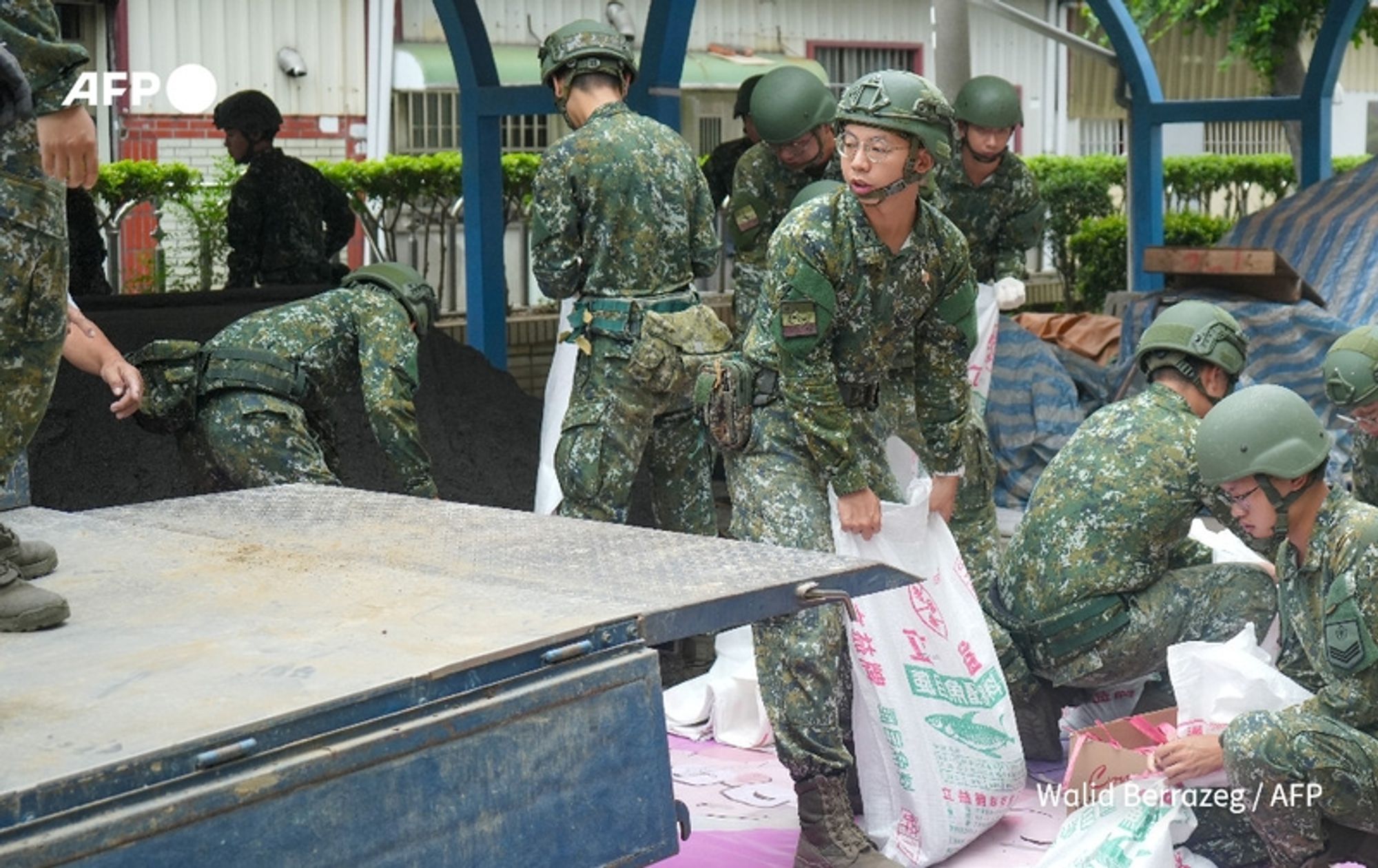 A group of soldiers in military fatigues and helmets is actively preparing for the incoming Typhoon Krathon in Taiwan. They are working in a partially covered area where bags of sand are being filled and stacked. The scene shows six soldiers engaged in the task; some are lifting heavy bags while others are positioned to receive or fill them. The environment appears to be an outdoor setting with a blue structure overhead providing some cover. In the background, there are hints of urban infrastructure, such as a building with windows and green foliage along the edges, indicating a city area. The atmosphere is focused and industrious, reflecting the urgency and seriousness of the task at hand as the typhoon approaches.