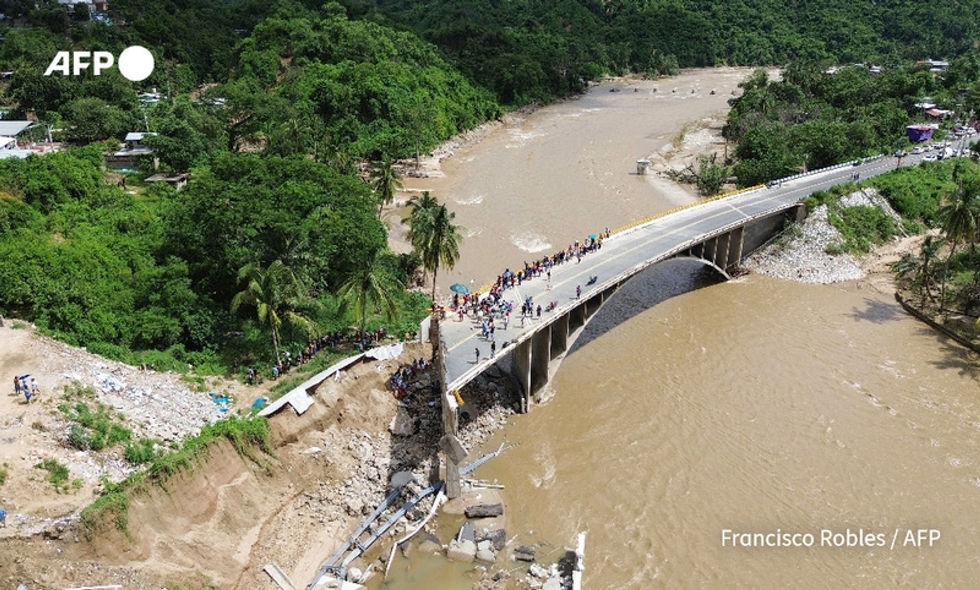 Aerial view of a damaged bridge located in a lush, green area following Hurricane John. The bridge appears partially collapsed on one side, with significant erosion visible on the riverbank. A group of people is gathered on the bridge, observing the damage and possibly in search of a way to cross. The river beneath is muddy and swollen, likely a result of the recent heavy rains from the hurricane. Surrounding the river are trees and dense vegetation, indicating a tropical environment. The image captures the devastation left by the storm, highlighting both the infrastructure damage and the response of the local community. In the top left corner, there is a logo of "AFP," and below it, the name "Francisco Robles / AFP" is displayed, indicating the source of the image.