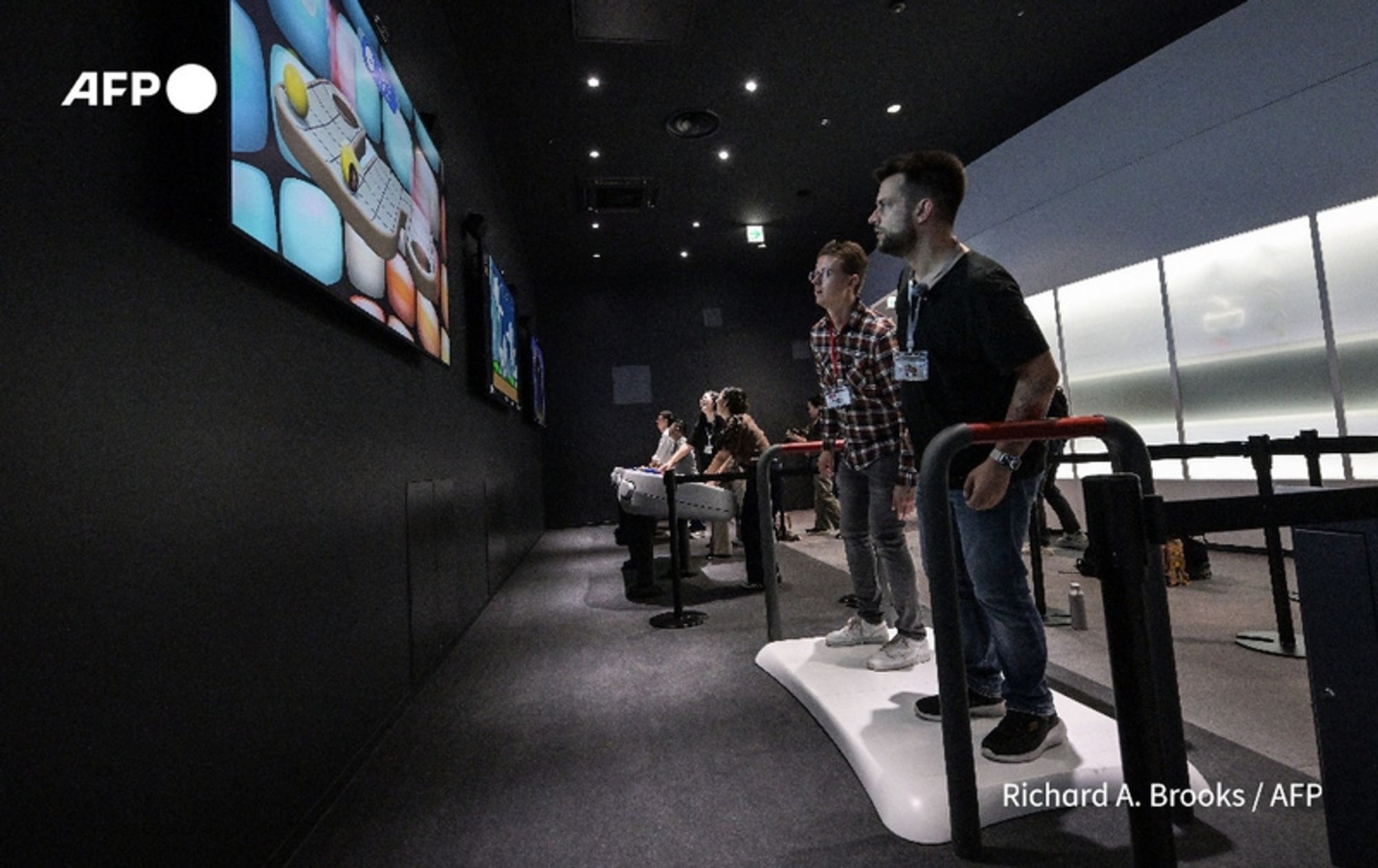 In a dimly lit exhibit hall within the newly opened Nintendo museum in Kyoto, several visitors interact with gaming displays. The scene is focused on a group of four individuals standing in front of large screens, engaged with the console games. Each person stands on a white interactive platform, designed for physical gaming, with their attention fixed on the colorful images projected on the screens. The screens display vibrant gameplay, likely showcasing Nintendo’s famous games. 

The first two individuals, positioned at the forefront, appear to be mid-action, smiling and enjoying their gaming experience. One person on the left wears a casual plaid shirt, while the person next to them is in a black t-shirt. Behind them, two additional visitors, a woman and a man, are also engaged with the games. The overall ambiance is lively, reflecting a sense of nostalgia and excitement for Nintendo’s extensive history in video gaming. The walls of the exhibition are adorned with subdued lighting, further enhancing the focus on the gamers and the vivid displays.