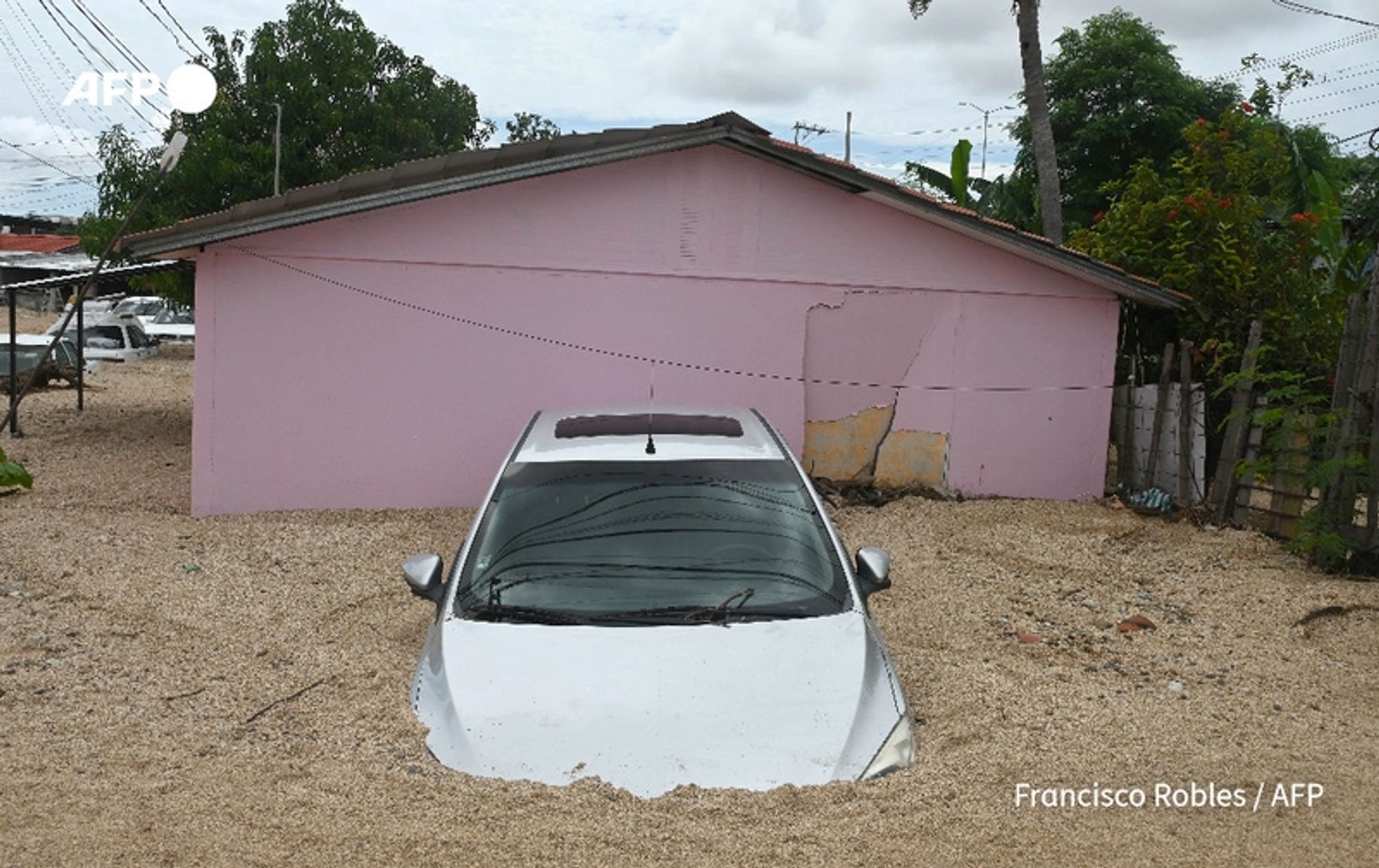 A white car is partially submerged in sandy debris in front of a pink house. The car's hood and windshield are visible above the sand, while the lower part is not seen. The pink house is in the background, showing some wear with patches and marks on the exterior walls. Surrounding the house are tall green trees and power lines, adding to the scene's context of a neighborhood impacted by a natural disaster. The sky is partly cloudy, indicating ongoing weather disturbances. The atmosphere conveys the aftermath of Hurricane John, reflecting the devastation and challenges faced by the local community.