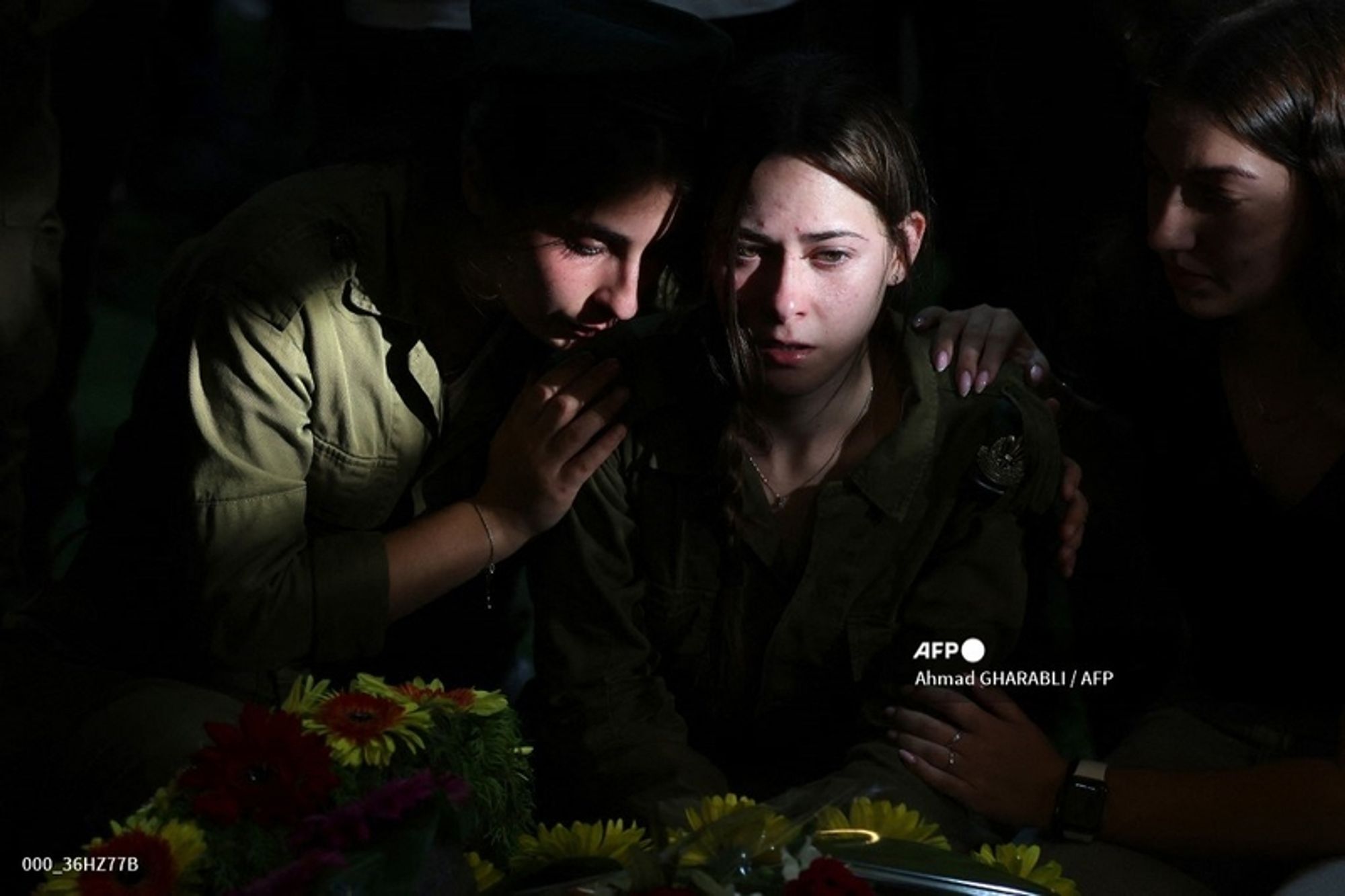 Two young women in military uniforms sit close together in a dimly lit setting. One woman, wearing a green military outfit, has her head bowed and appears visibly upset, with tears in her eyes. Her expression reflects deep sorrow and grief. The other woman, also in military attire, leans in closely, providing comfort and support by placing her arm around the distressed individual. They are surrounded by a small arrangement of colorful flowers that add a touch of brightness to the scene. Additional figures can be seen in the background, though they are slightly out of focus, contributing to the somber atmosphere. The image captures a poignant moment of emotional solidarity in a time of loss.