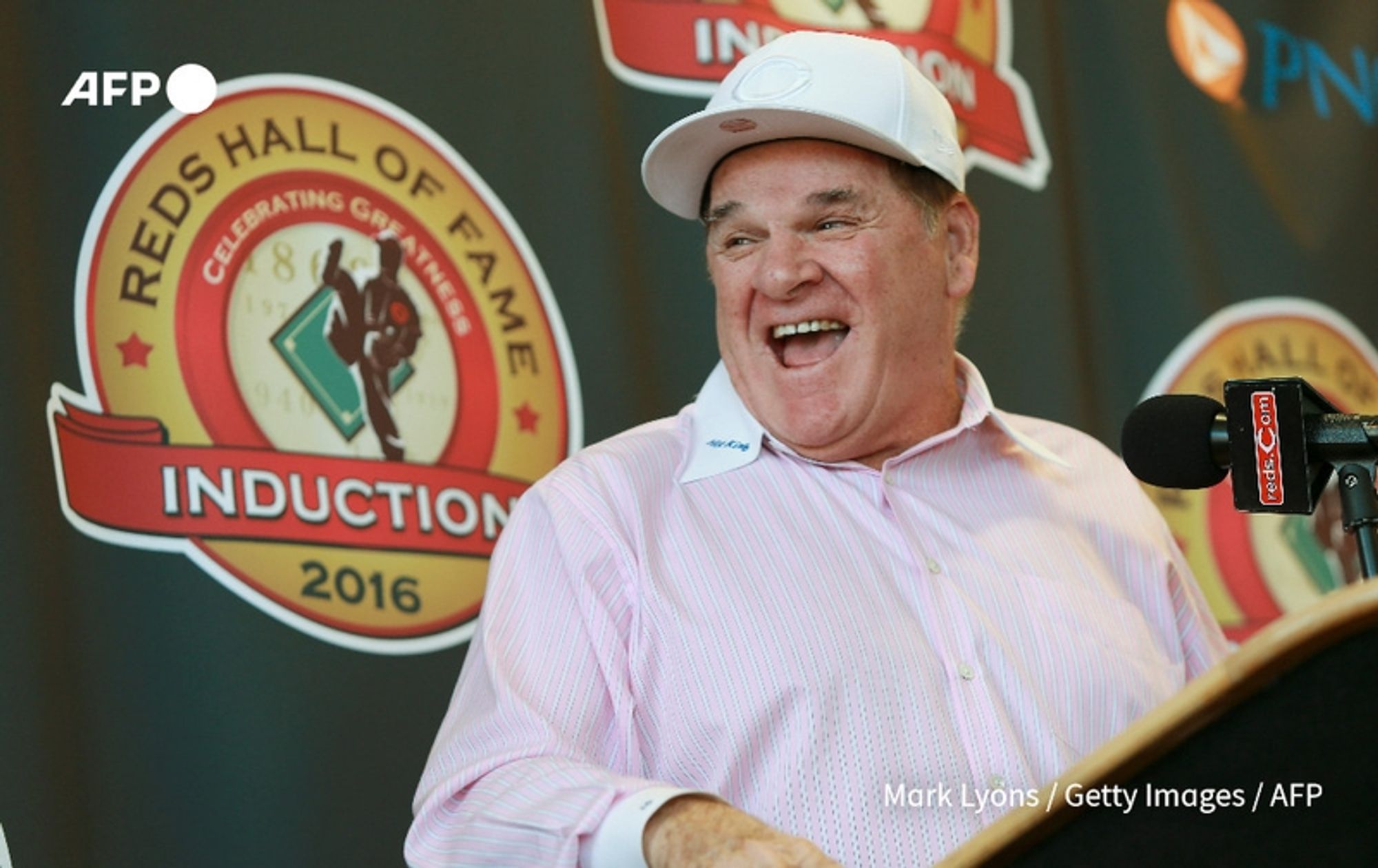 A middle-aged man with a joyful expression is standing at a podium, smiling broadly. He has short hair and is dressed in a light pink collared shirt, which has a subtle pattern, and a white baseball cap. The background features a large circular emblem that reads "REDS HALL OF FAME" in bold red letters, with the words "INDUCTION" prominently displayed. The emblem includes various baseball motifs and the year "2016." The setting suggests a celebratory event related to the Cincinnati Reds, likely an induction ceremony. The man's demeanor conveys happiness and warmth, suggesting a positive moment during the event.