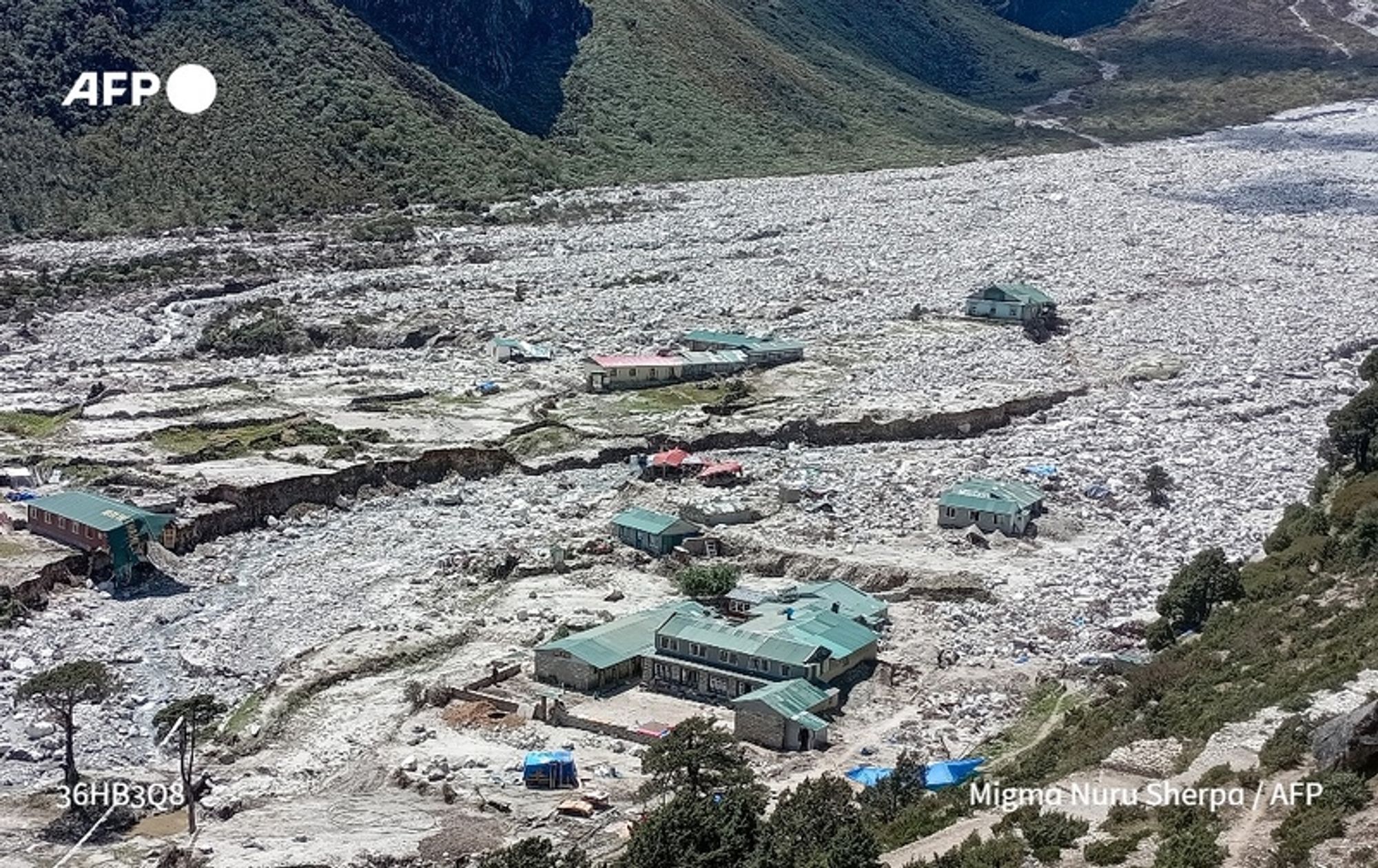 A wide aerial view of the village of Thame, located in the foothills of Mount Everest, showing extensive damage caused by flooding from a glacial lake burst. The landscape is dominated by a massive expanse of white ice and debris, covering the ground in a chaotic pattern. Several buildings can be seen scattered throughout the image, with some structures partially buried or isolated by the surrounding snow and ice. The buildings have green roofs and are painted in various colors, including red and cream. Alongside the buildings, patches of greenery and trees are visible, contrasting with the icy floodwaters. The scene captures the aftermath of the disaster, emphasizing the scale of the flooding and the impact on the village's infrastructure, reflecting concerns about the effects of climate change in the Himalayan region.