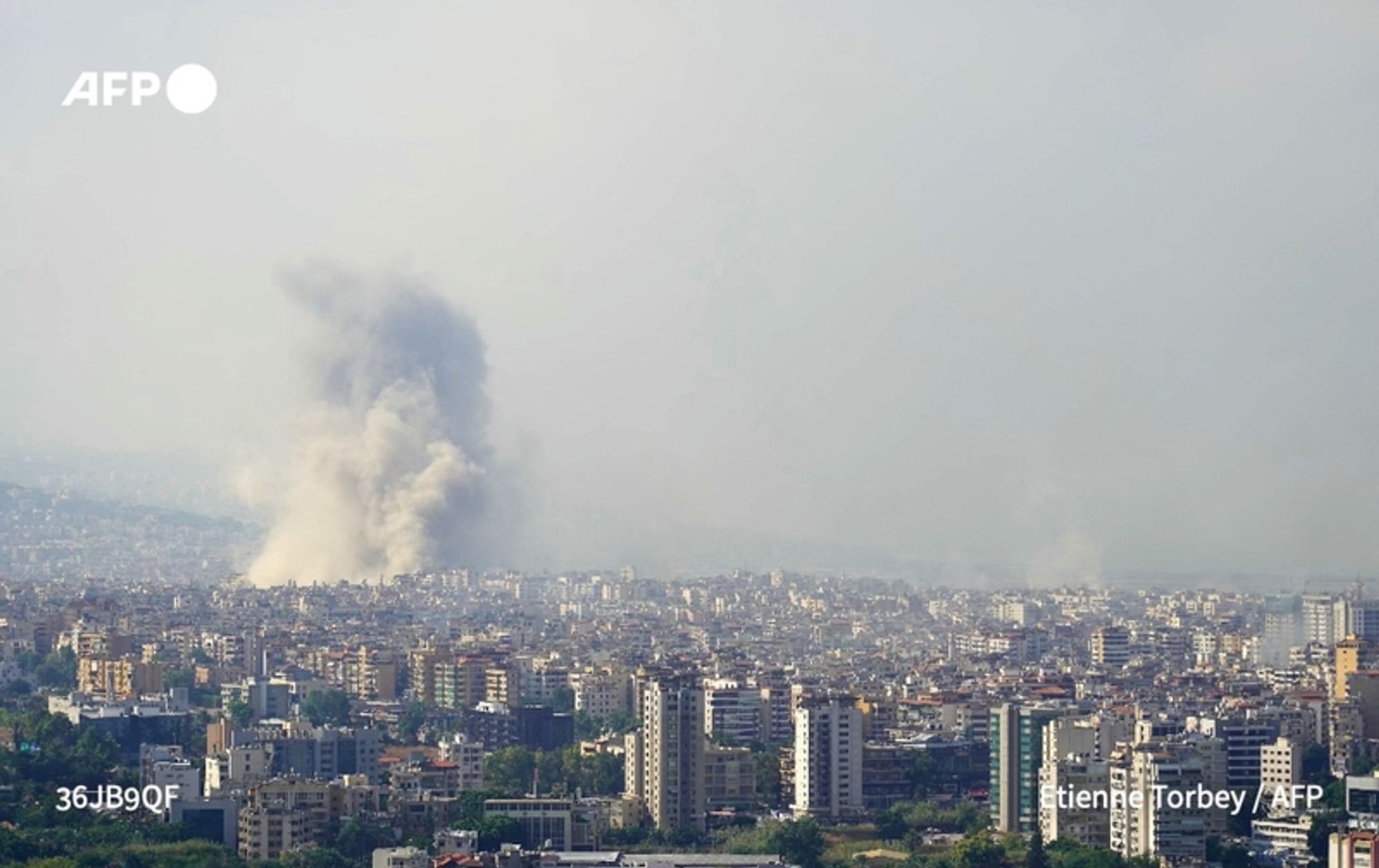 A panoramic view of a cityscape in Beirut is captured, dominated by a large column of gray smoke rising into the sky, indicating a recent explosion or fire. The smoke billows up from a location not visible in the image. The foreground displays densely packed buildings, showcasing a mix of architectural styles, with several high-rise structures and mid-sized apartment complexes visible. The overall atmosphere appears tense, with the sky hazy, suggesting chronic disturbances in the area. The backdrop features a mix of urban and mountainous geography, reflecting the landscape's natural beauty juxtaposed against the turmoil occurring. The image paints a vivid picture of the impact and aftermath of military actions in the region.