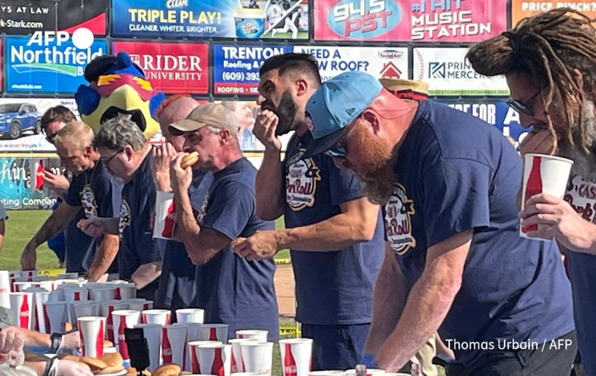 A group of male competitors, dressed in matching dark blue t-shirts with a logo that reads "Pork Roll," are engaged in a competitive eating contest. Each contestant is positioned at a long table filled with numerous pork rolls and drinks in styrofoam cups. The atmosphere is lively, with colorful event signage in the background promoting local businesses and sponsors. Some competitors are in the act of eating, focusing intently on their rolls, while others appear to pause, possibly strategizing for the next bite. A mascot, resembling a large bird, is visible in the background, adding to the festive ambiance. The bright blue sky and the green grass of the field suggest a sunny day, typical for an outdoor event. The scene captures the excitement and camaraderie of this very American pastime of competitive eating.