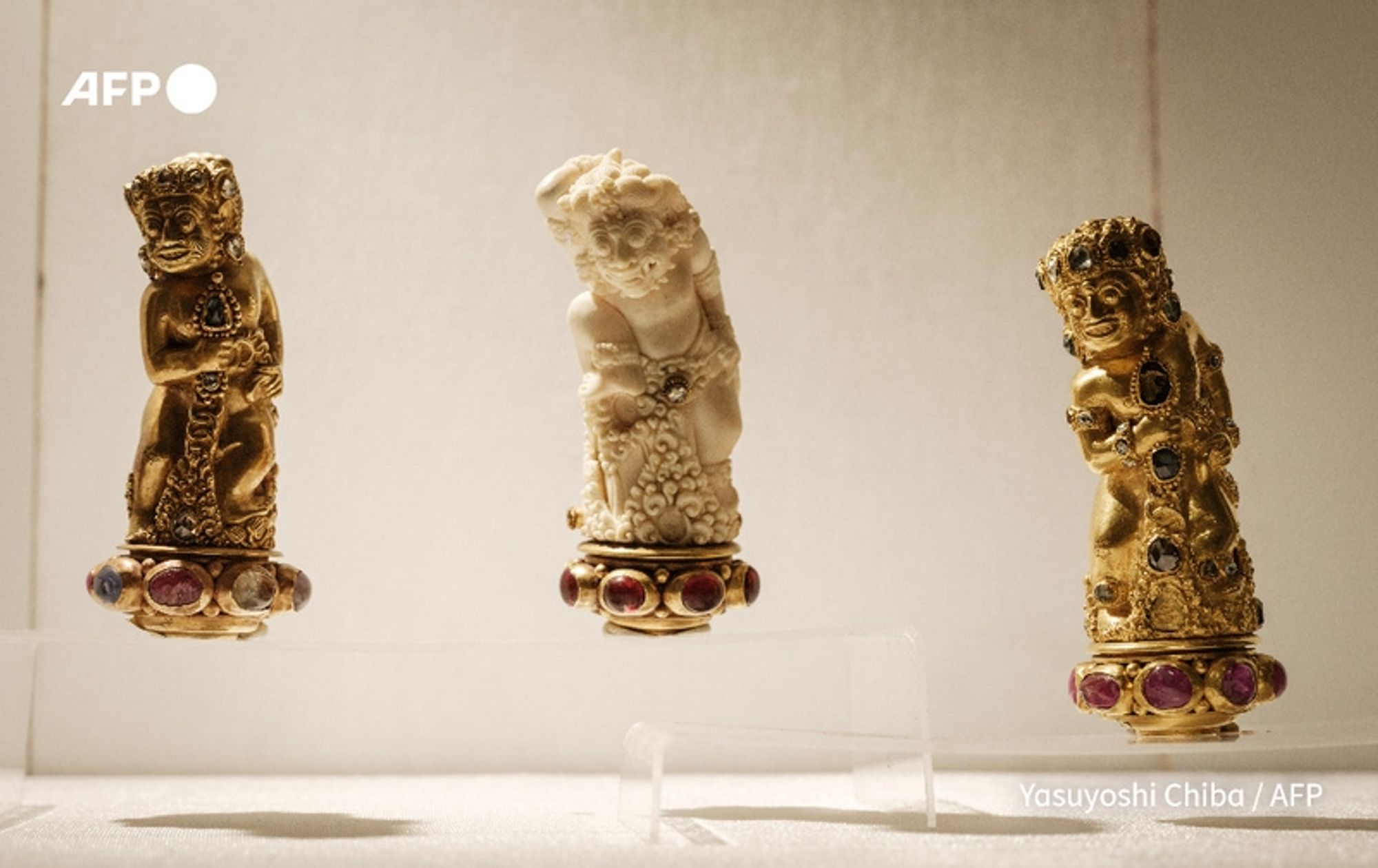 Three exquisite jewelry artifacts are displayed on a white pedestal at the National Museum of Indonesia in Jakarta. The centerpiece is a finely crafted statue of Brahma, depicted in a gold and jeweled form, adorned with multicolored gemstones, showcasing intricate detailing. Flanking Brahma, two additional smaller figures are present; one is a golden figure with elaborate decorations and a similar collection of gemstones, and the other is a white sculpture, elegantly carved to portray a graceful posture. The pieces highlight the richness of Indonesian artistry and craftsmanship, symbolizing a significant cultural heritage. The backdrop is minimalistic, drawing attention to the artifacts themselves, which are part of the repatriated Lombok collection.