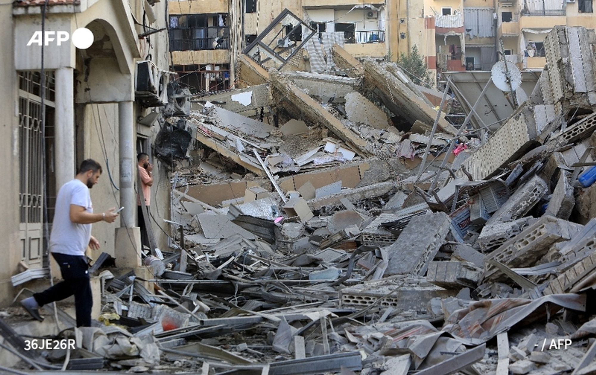 A person walks through rubble on a street in Beirut, surrounded by the destruction of collapsed buildings. The debris is scattered across the ground, consisting of concrete blocks, metal scraps, and building materials. Visible remnants of walls and windows indicate recent destruction, and several nearby structures exhibit signs of damage, with broken glass and exposed frameworks. In the background, a man is seen standing among the ruins, while another individual appears to be on a mobile device. The overall scene conveys a sense of chaos and devastation, reflecting the aftermath of recent intense strikes in the area.