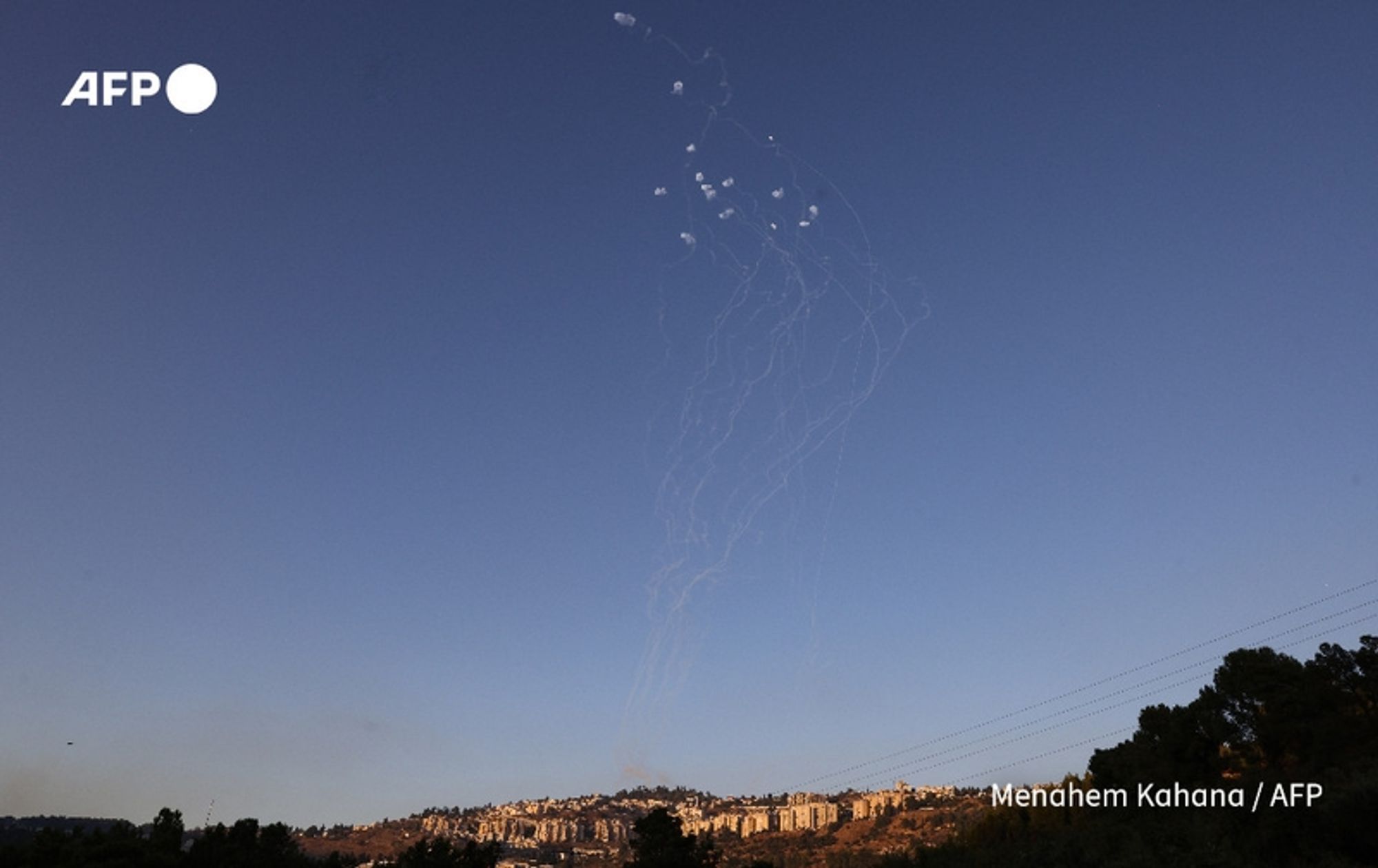 A view of the sky during an active conflict, featuring several white smoke trails ascending into a clear blue sky. The trails are emanating from a point on the ground and appear to be a result of rockets or projectiles being launched. In the background, a hilly landscape is visible, with a residential area marked by a series of buildings and houses atop a hill. The scene is set against a serene atmosphere despite the military activity, highlighting a stark contrast between the natural environment and the ongoing tension. The image captures the ongoing hostilities, reflecting the current conflict involving Hezbollah militants in Lebanon.